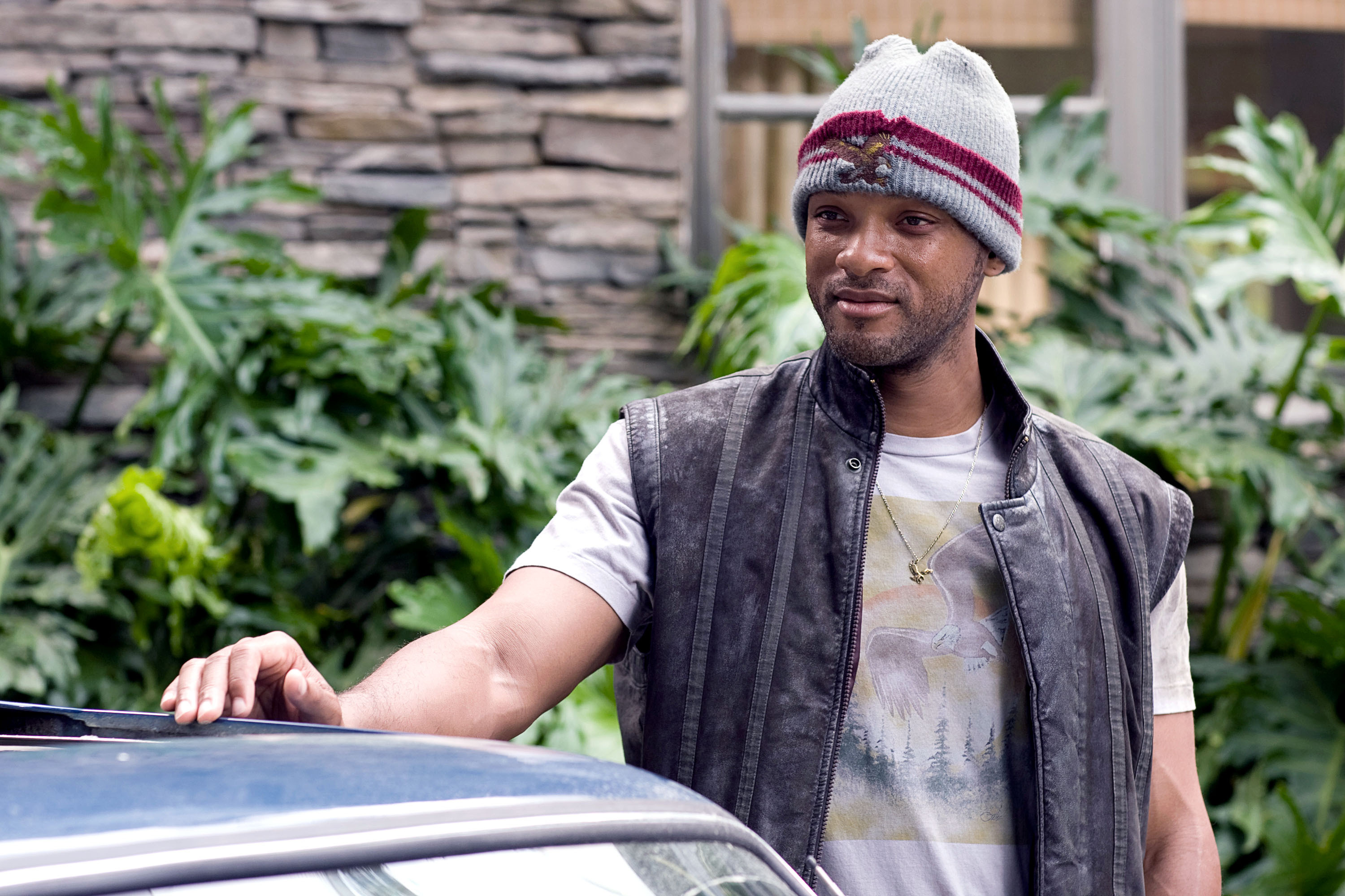 Will Smith wearing a casual outfit with a beanie and vest, resting his hand on a car in an outdoor setting in a scene from &quot;Hancock&quot;