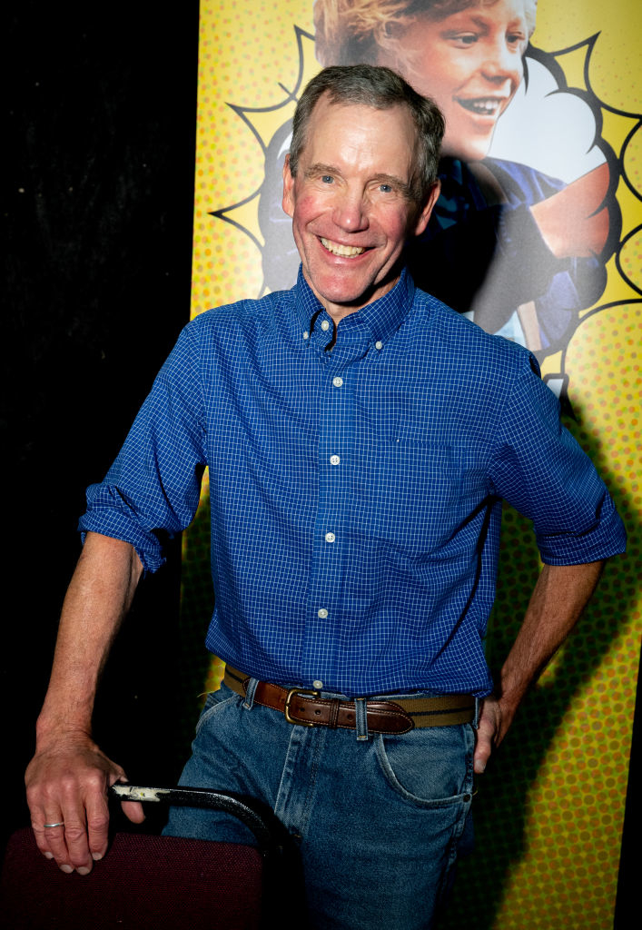 A man in casual attire, denim jeans, and a blue shirt, stands smiling with one hand on a chair in front of a colorful poster backdrop featuring a laughing child