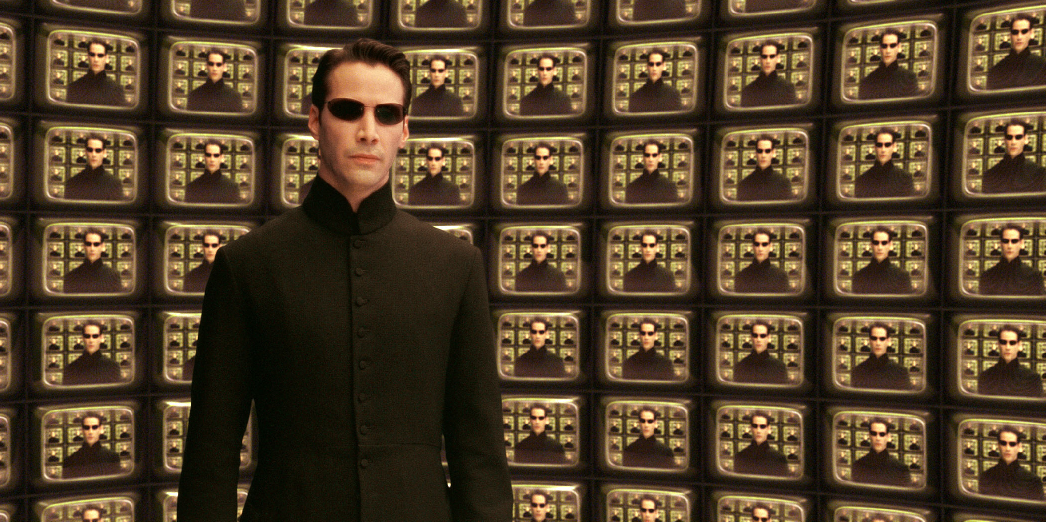 Keanu Reeves as Neo, wearing a sleek black outfit and dark sunglasses, stands in front of a TV with a mirror image of himself in a scene from &quot;The Matrix Reloaded&quot;
