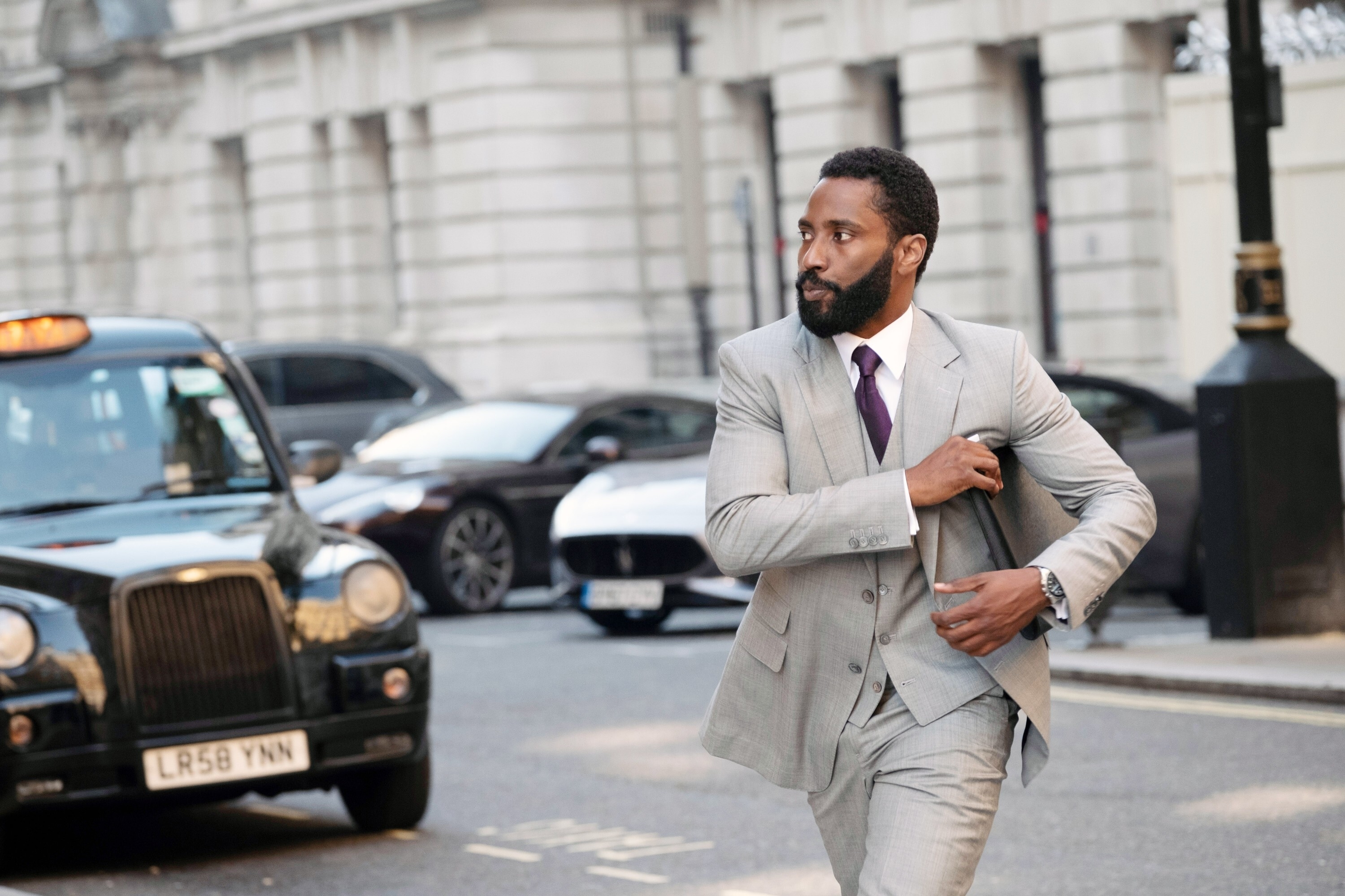 John David Washington in a gray suit and tie, walking on a city street, seemingly adjusting his jacket. Cars and a taxi are visible in the background in a scene from &quot;Tenet&quot;