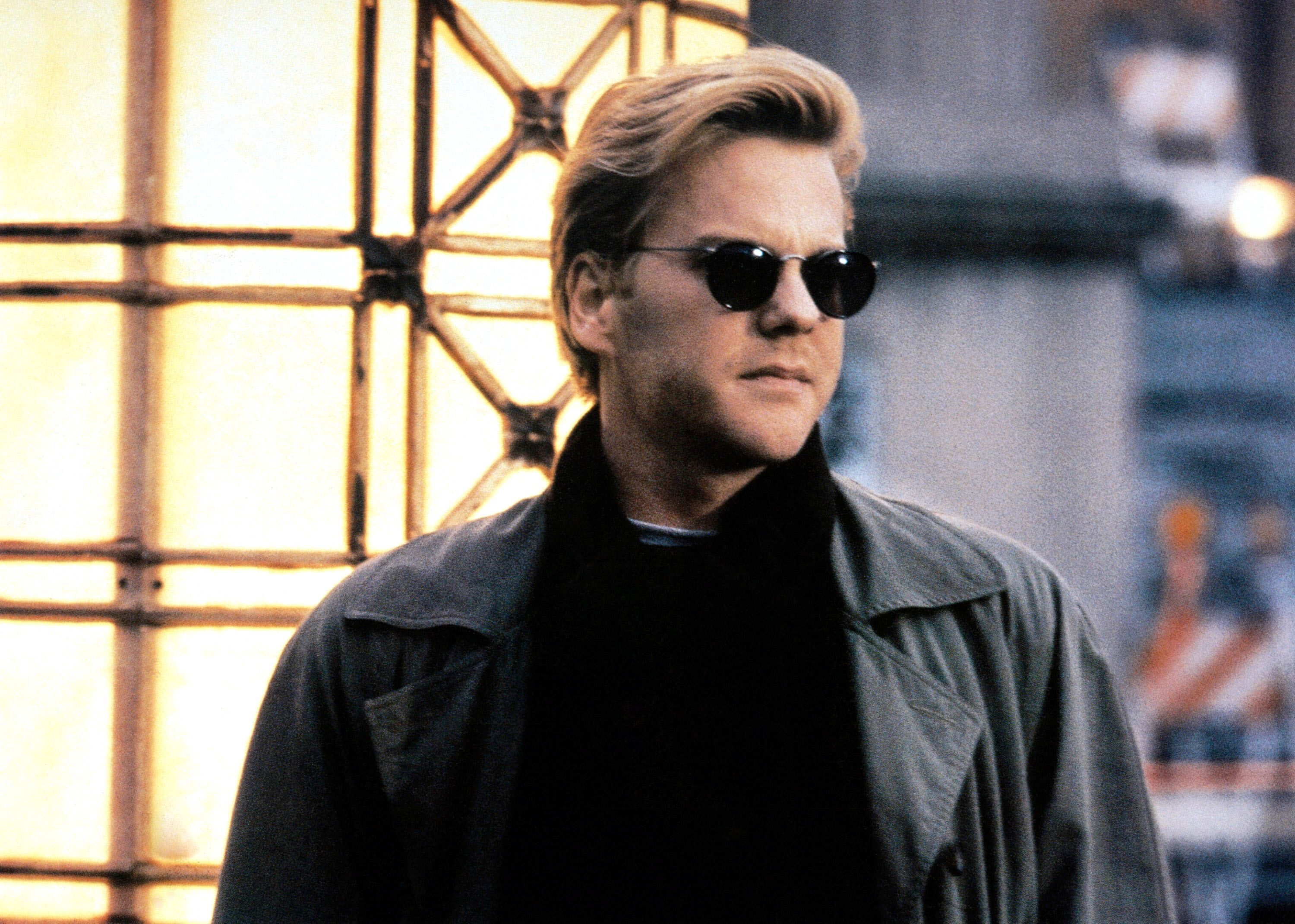 Kiefer Sutherland, wearing a trench coat and dark sunglasses, stands in front of a lit structure in a scene from &quot;Flatliners&quot;