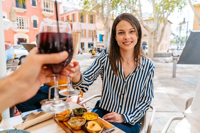 A woman in a striped blouse enjoys a meal and raises a glass of wine in a toast at an outdoor restaurant. Another person&#x27;s hand holding a glass is in the foreground