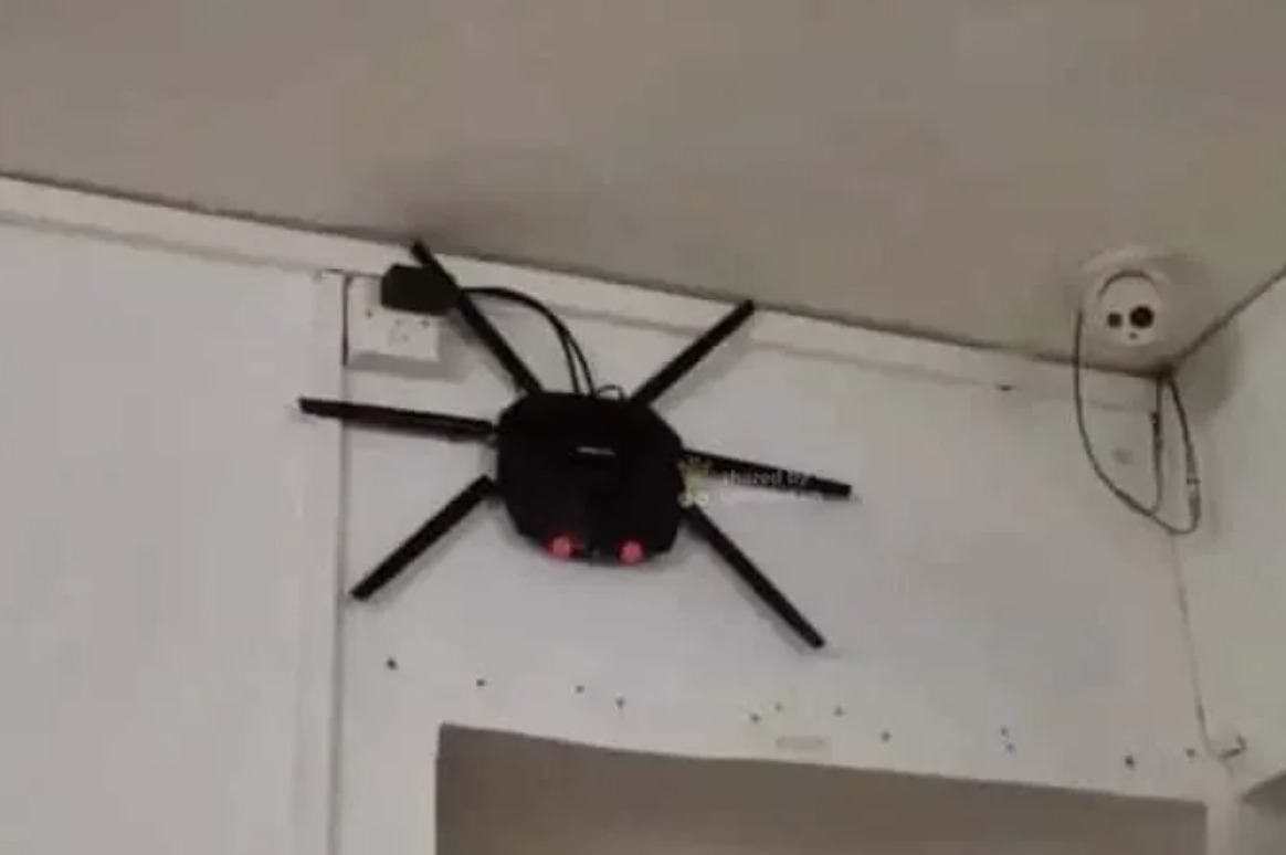 A Wi-Fi router installed high on a wall is humorously modified to resemble a spider with added legs. A security camera is mounted nearby