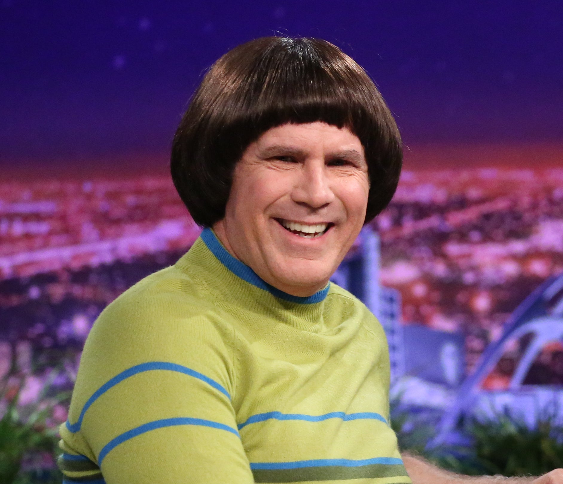 Will Ferrell, dressed in a green striped sweater and white pants, sits on a talk show set, smiling and wearing a bob wig