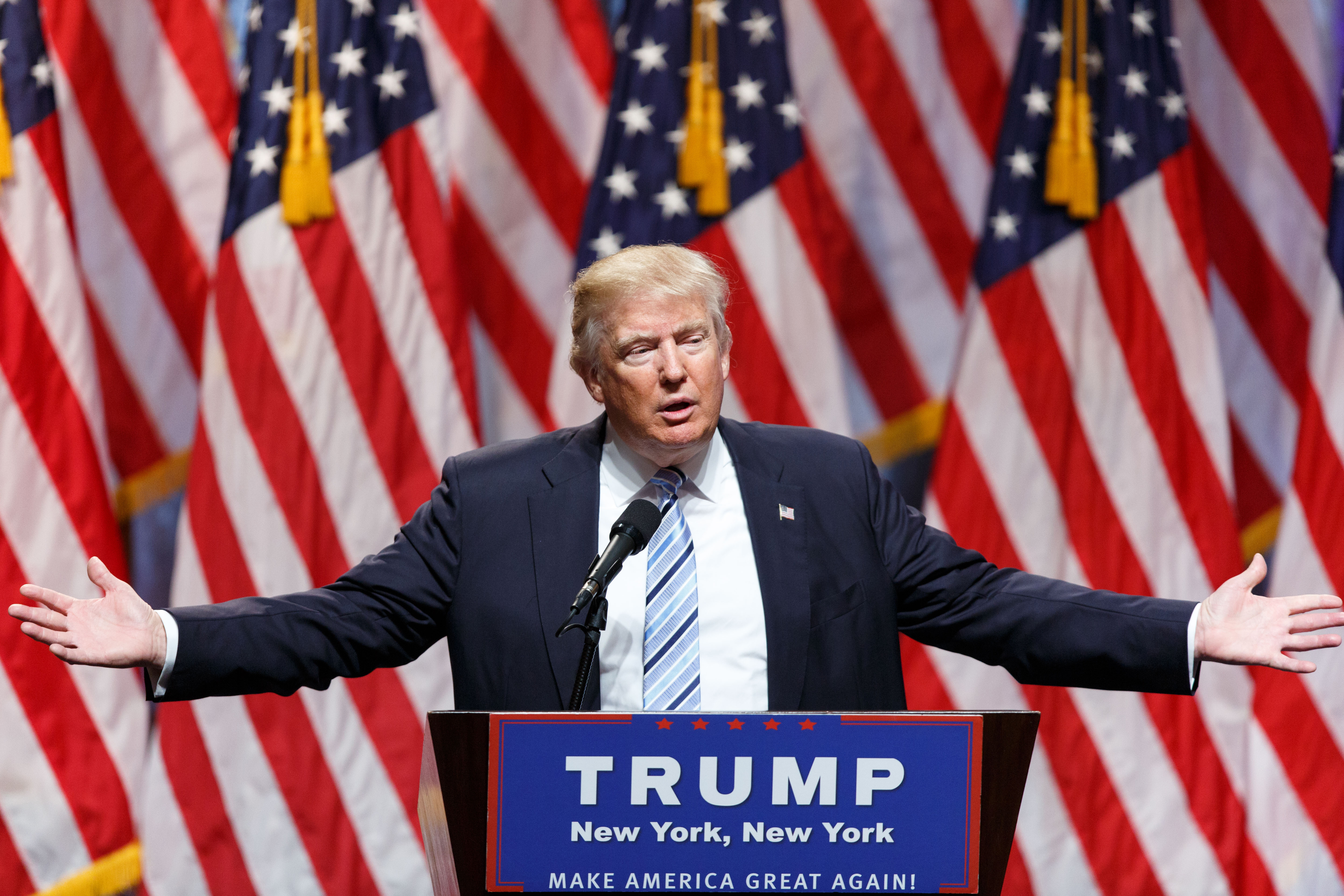 Donald Trump speaking at a podium with arms outstretched, American flags in background. Podium says &quot;TRUMP New York, New York. Make America Great Again!&quot;