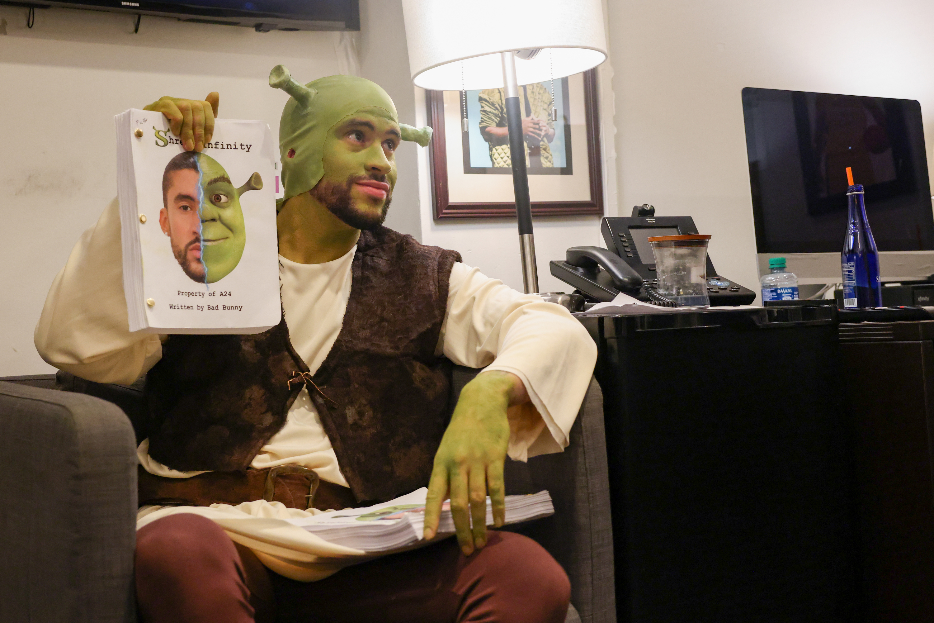 Bad Bunny dressed as Shrek sits holding up a printed picture divided into his face and Shrek's