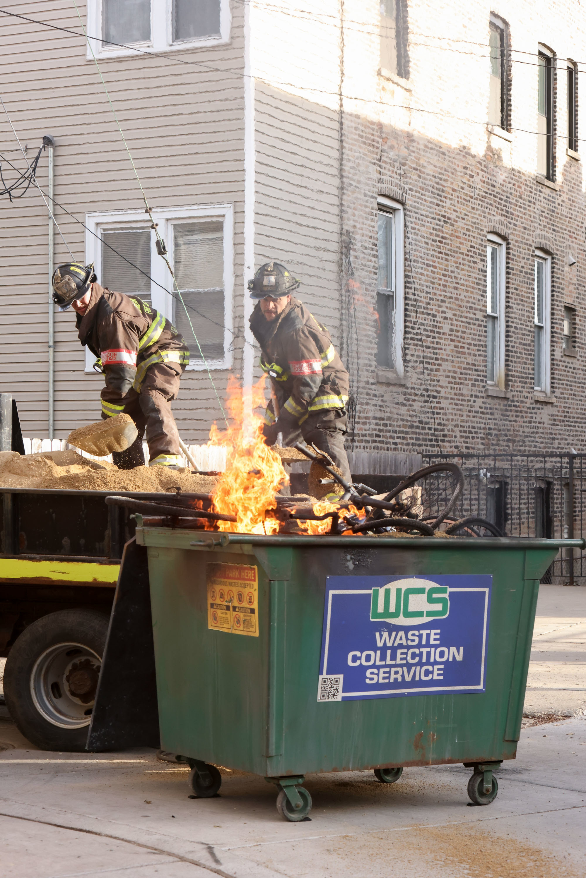 Two firefighters extinguish a fire in a green Waste Collection Service dumpster in an urban alley. Flames and smoke are visible