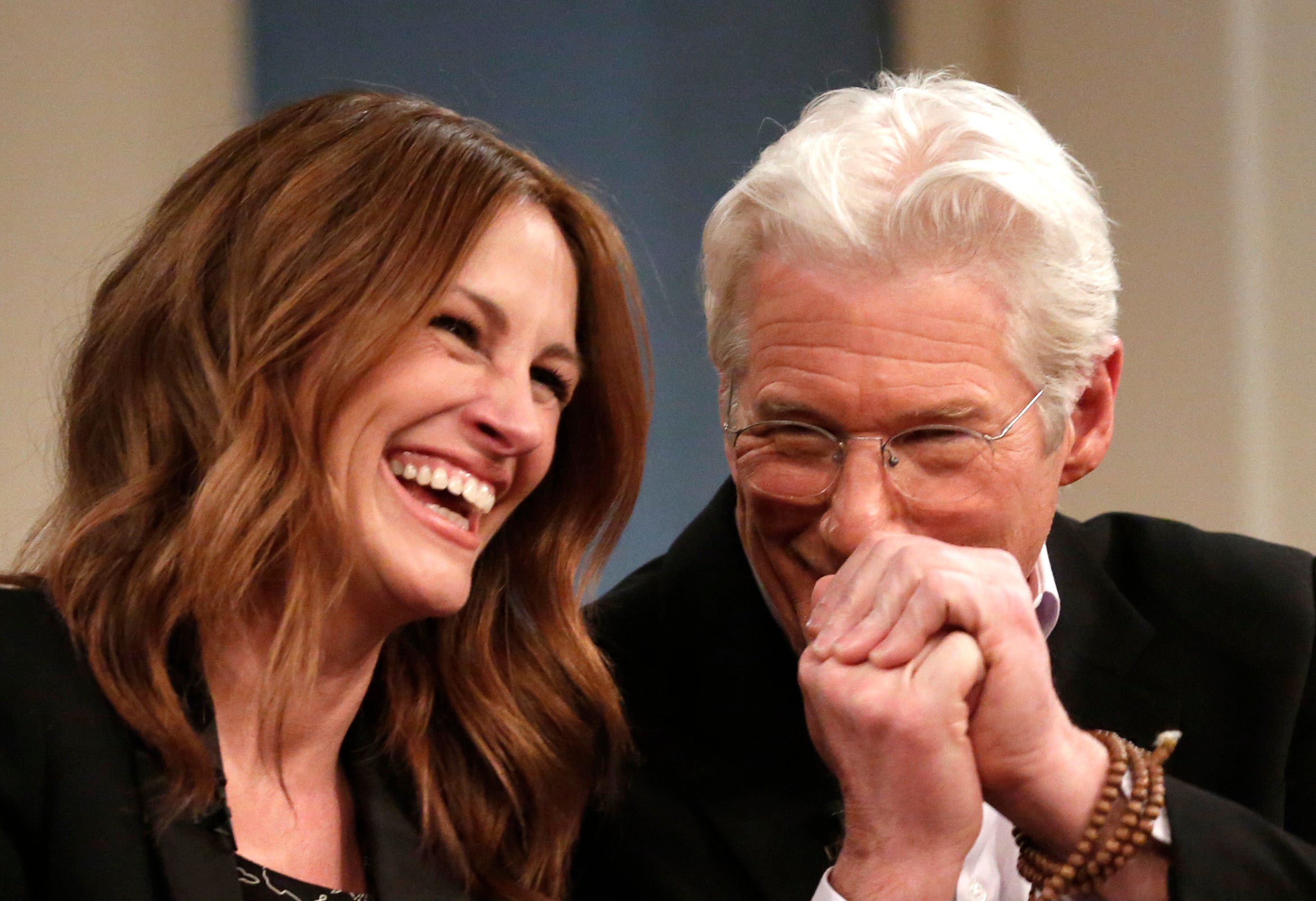 Julia Roberts and Richard Gere, both smiling and laughing, are seated holding hands during an interview. Julia wears a suit jacket over a patterned top