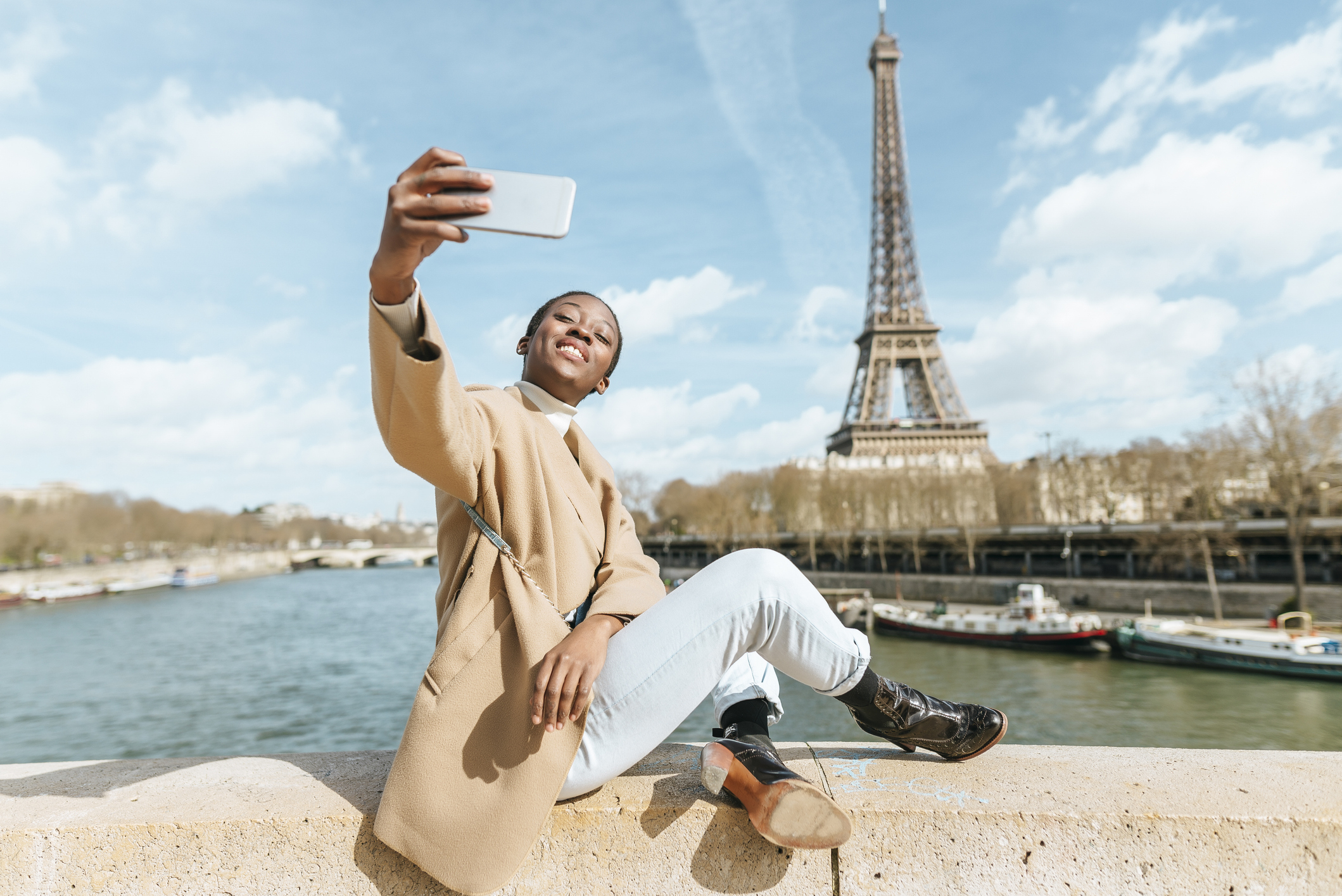 A person takes a selfie while sitting on a ledge by the Eiffel Tower. They wear a light coat, white pants, and black shoes. The sky is partly cloudy
