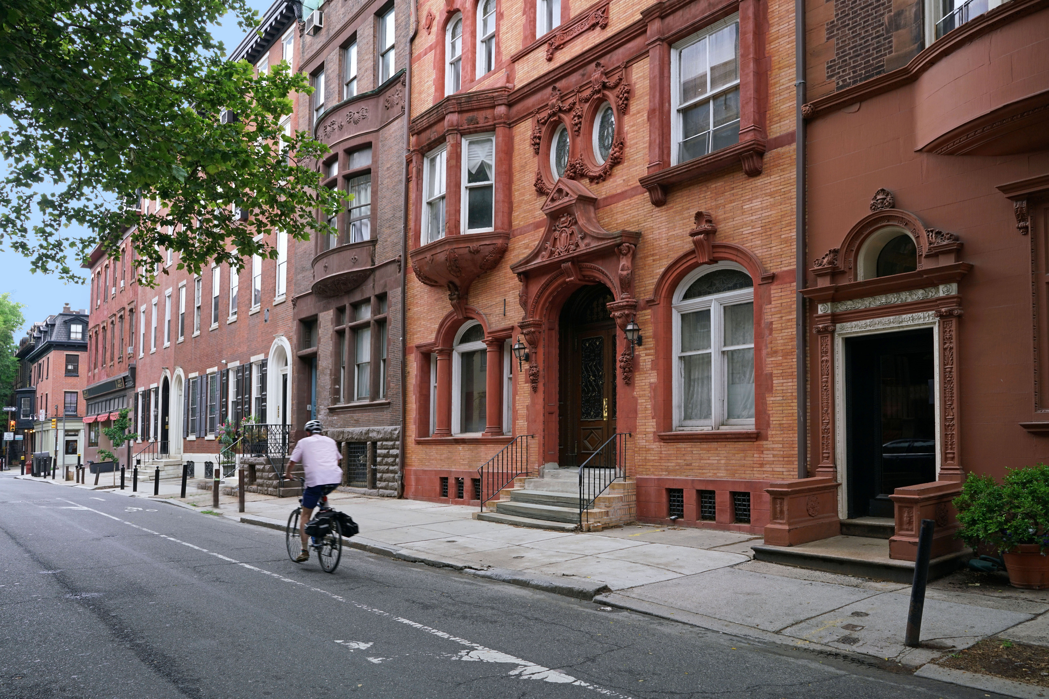 A cyclist in a white shirt rides down a quiet residential street lined with historic brownstone buildings