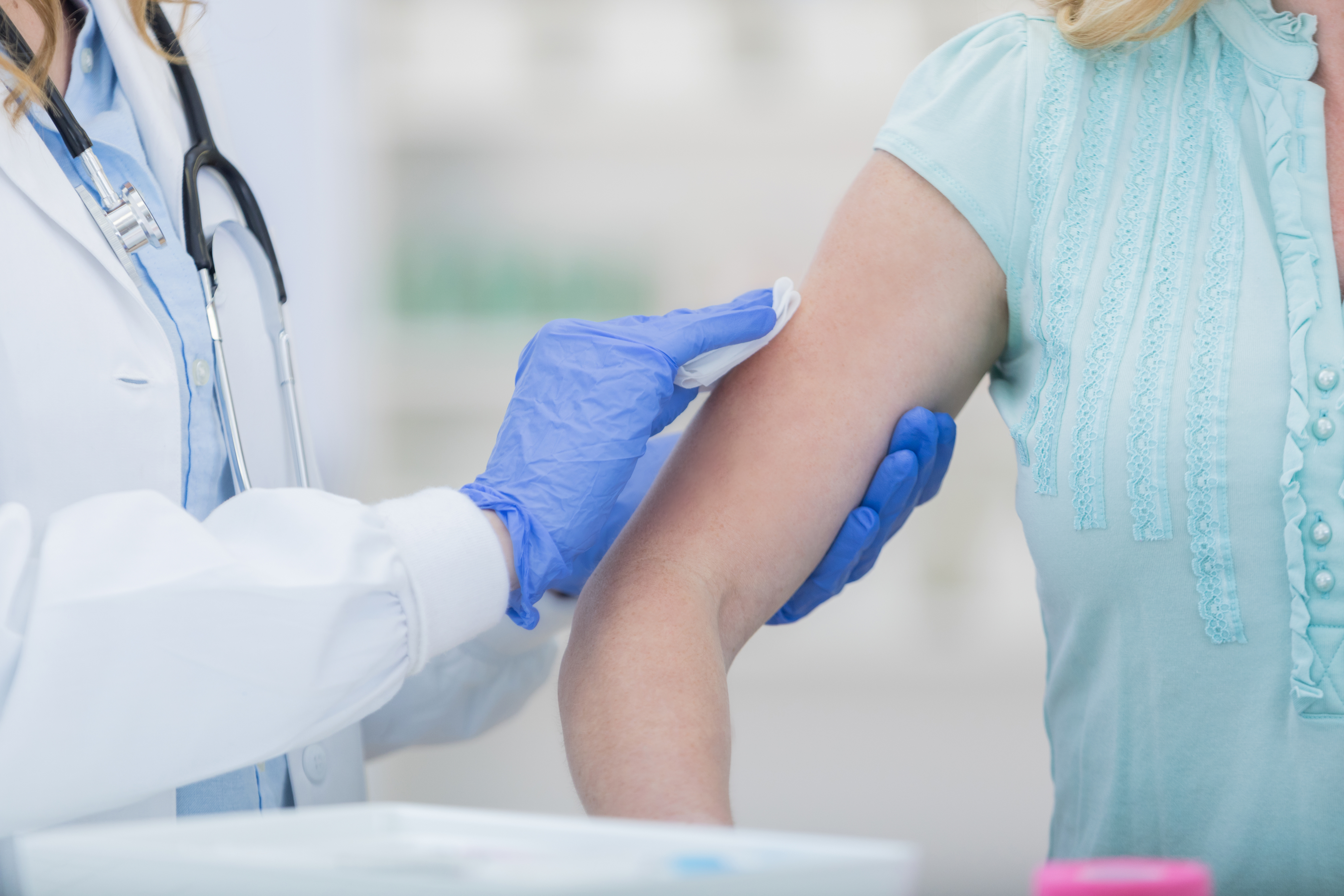 A healthcare professional in a white coat and blue gloves disinfects a patient's arm, preparing to administer a vaccine