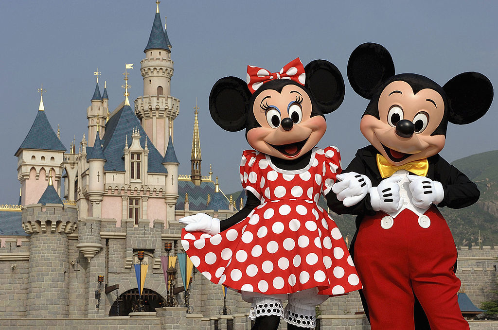 Minnie Mouse in a polka-dot dress and Mickey Mouse in red shorts and yellow shoes stand happily in front of a castle at Disneyland