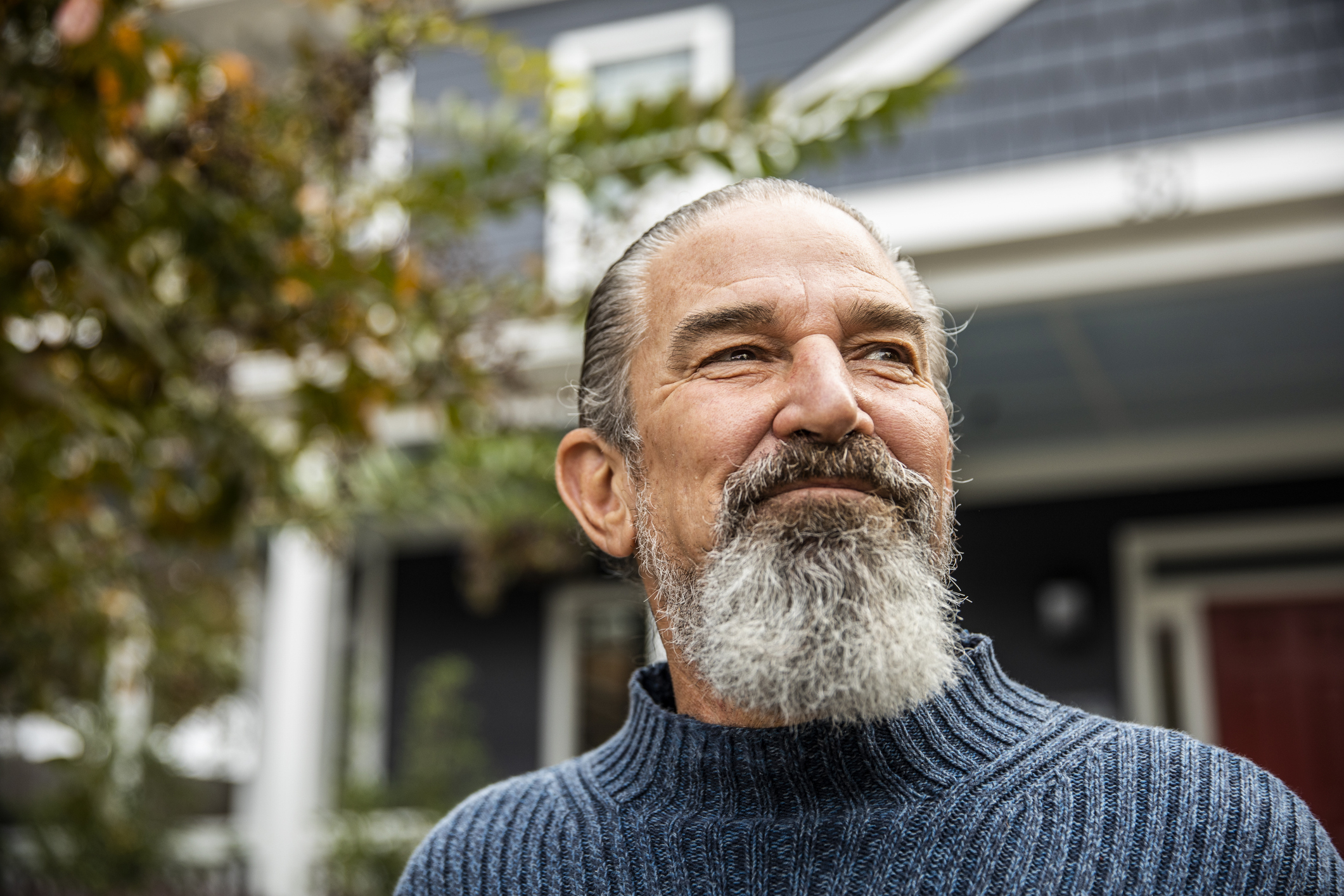 A man with a gray beard and pulled-back hair, wearing a ribbed turtleneck sweater, stands outside a house and looks off into the distance with a thoughtful expression