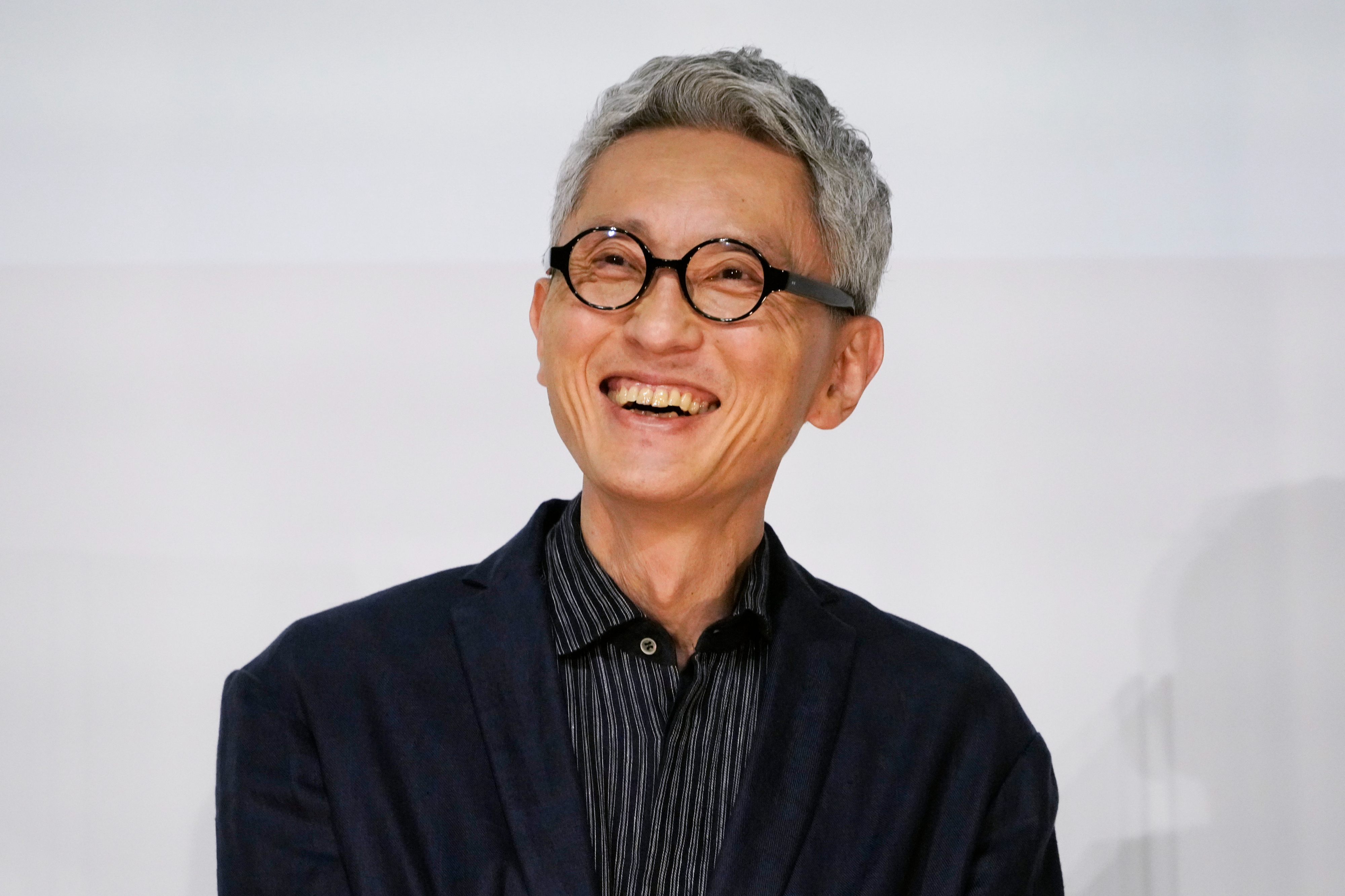 男性が笑顔でカメラを見ています。彼はメガネをかけ、ジャケットとシャツを着用しています。