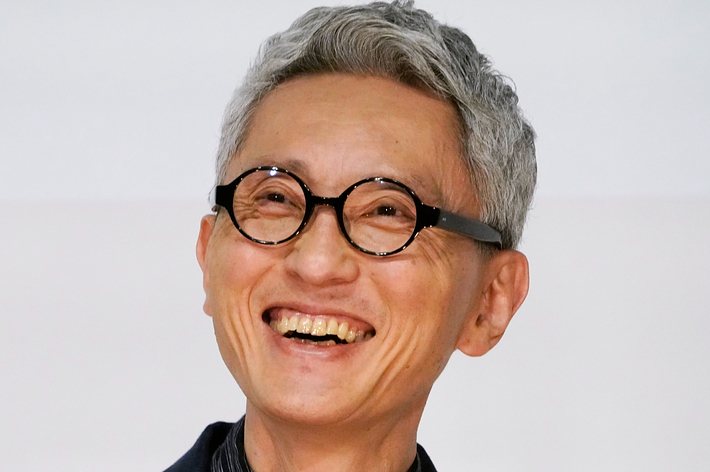 男性が笑顔でカメラを見ています。彼はメガネをかけ、ジャケットとシャツを着用しています。