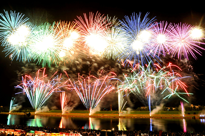 夜空に大規模な花火が打ち上げられ、多くの人々が川沿いで観賞しています。