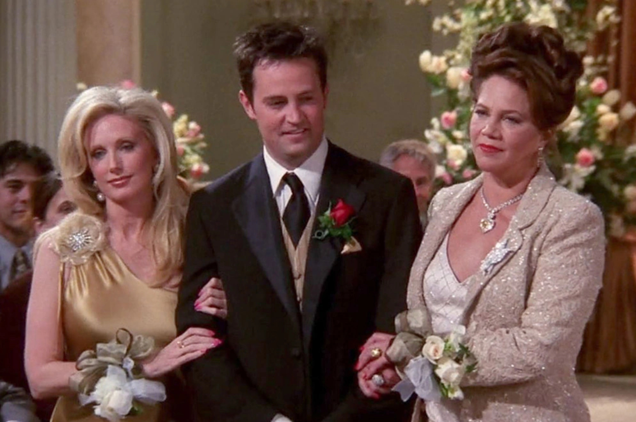 Matthew Perry in a black suit with Morgan Fairchild and Kathleen Turner in elegant gowns, all standing together at an indoor formal event with flowers in the background
