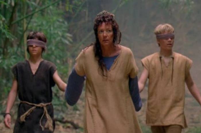 Three blindfolded individuals, led by a woman, walk through a forest. They wear simple, rustic clothing