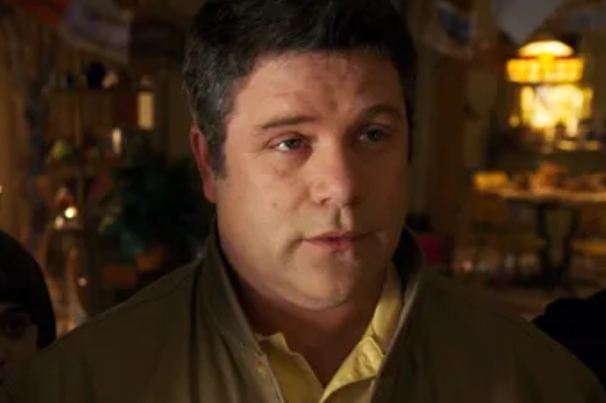 Man standing indoors with a neutral expression, wearing a light-colored shirt under a brown jacket. The background is softly blurred with warm lighting