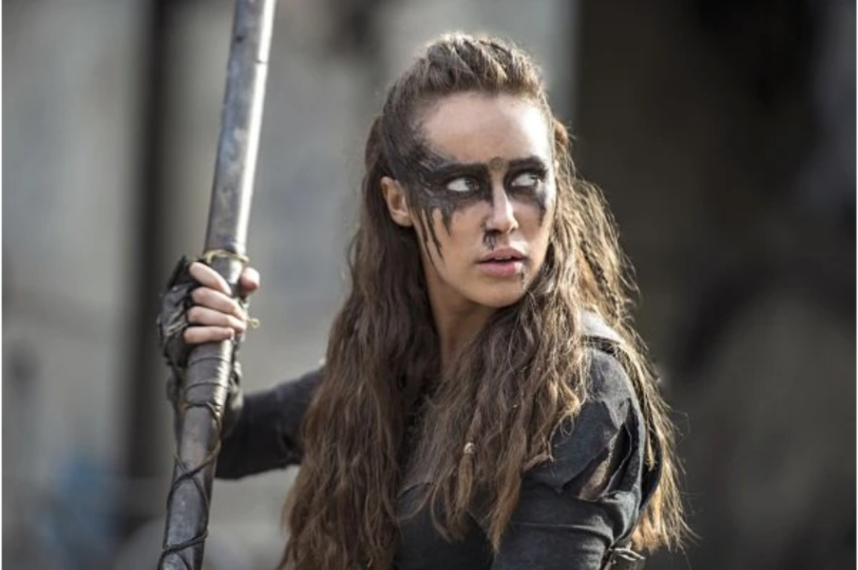 Alycia Debnam-Carey dressed as a warrior, holding a spear and with dramatic face paint, in a determined pose from her role in the series "The 100."