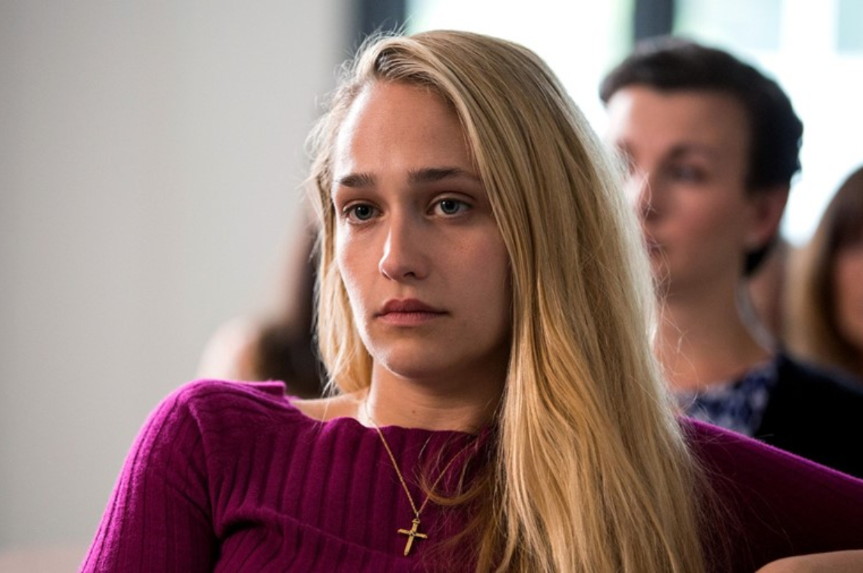 Jemima Kirke is sitting indoors, wearing a simple top and a cross necklace. She has a serious expression and her hair is down