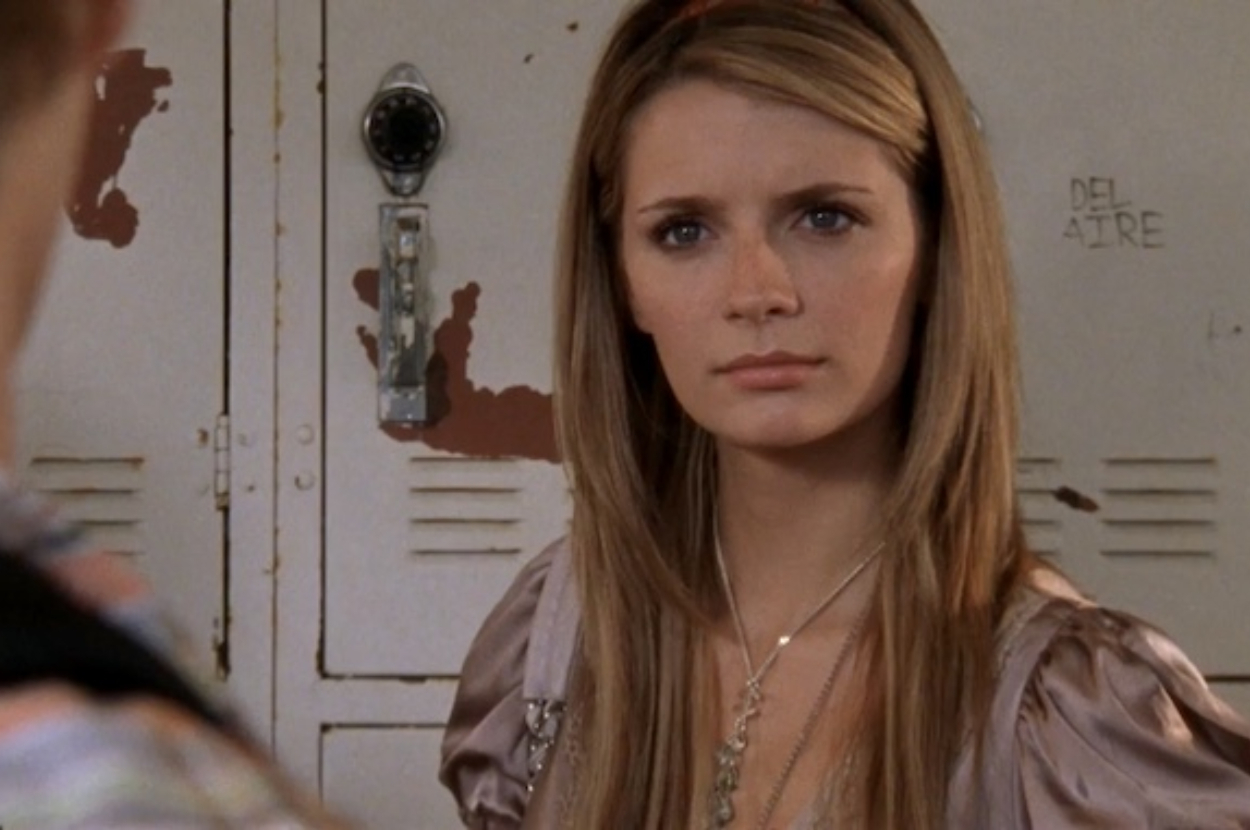 Mischa Barton standing in front of lockers, looking serious with long, straight hair and a pendant necklace