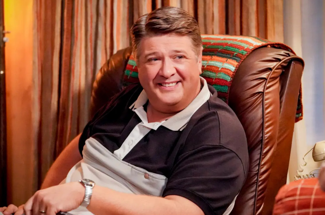 Man sitting comfortably in a chair, smiling while looking to his side. He is wearing a collared shirt with black and white sections and a wristwatch