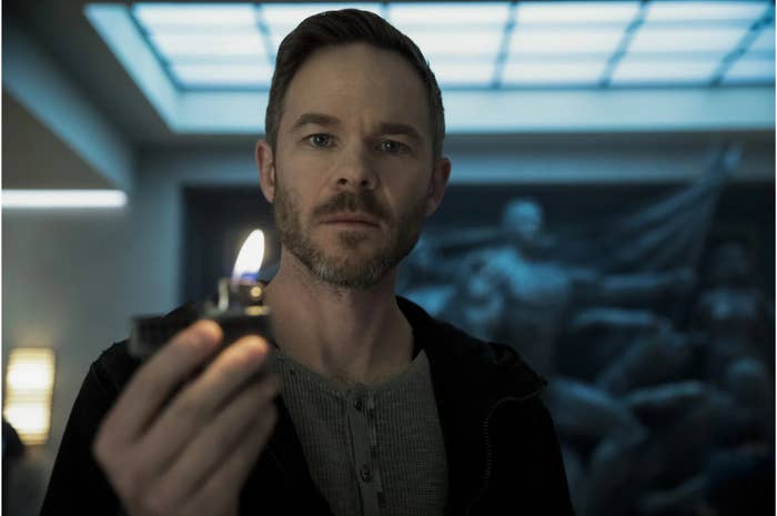 A bearded man with short hair holds a lit lighter in a dimly lit room with statues in the background