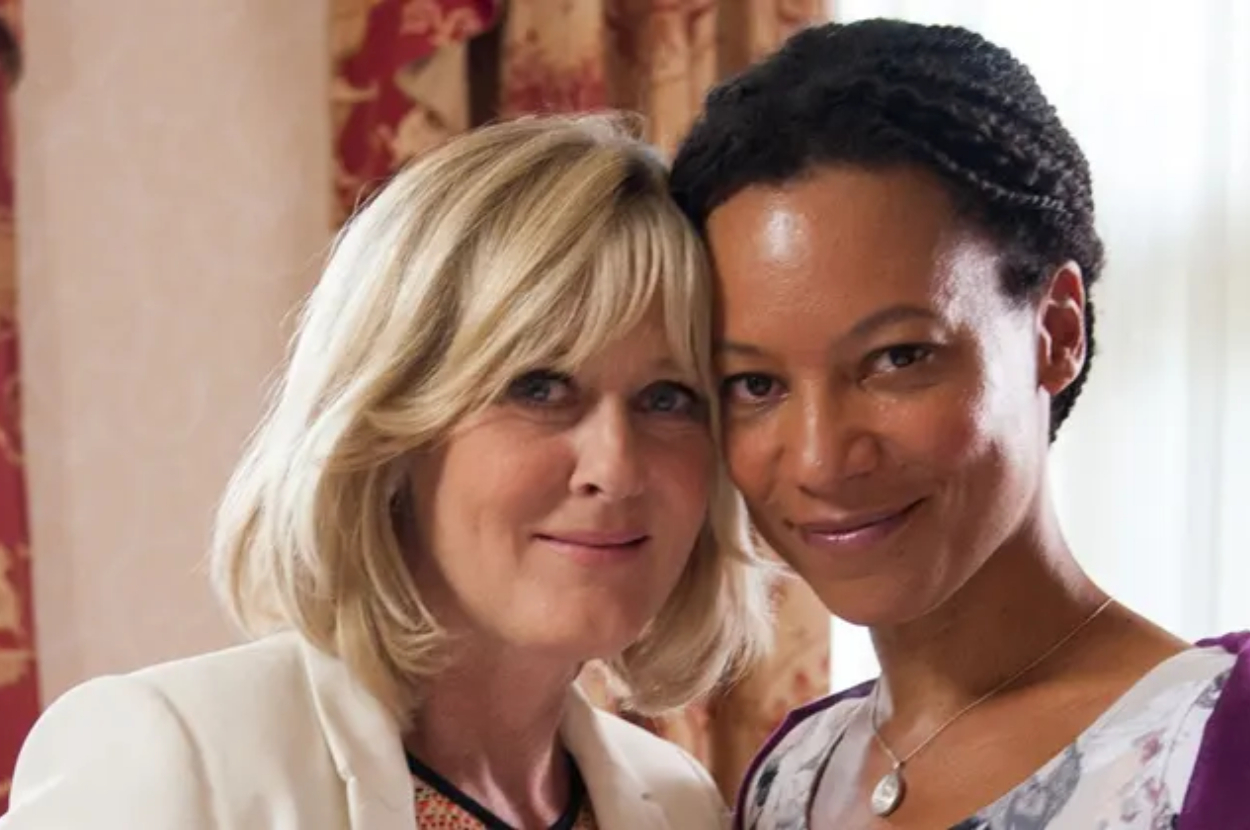 Two women, one with short blonde hair in light-colored clothing, the other with braided hair and a purple top, smile together closely
