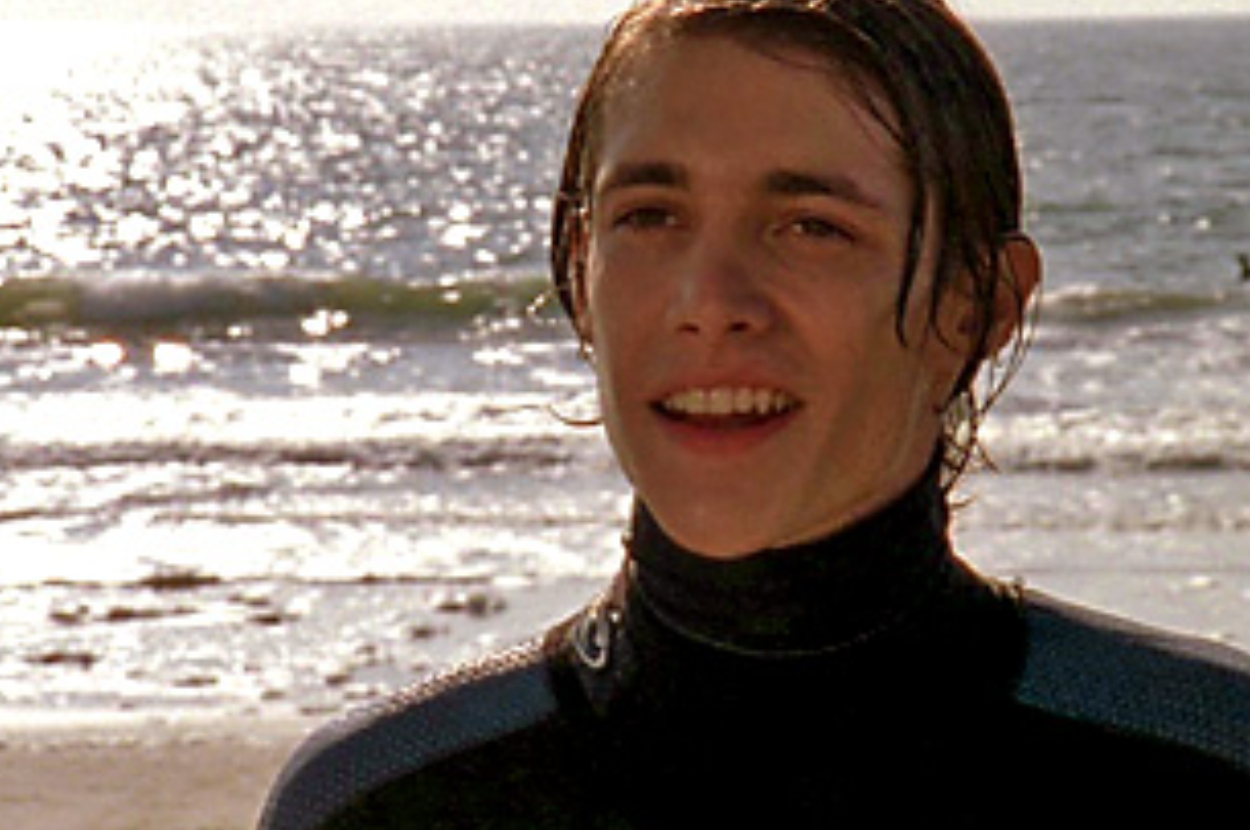 A young man stands on a beach, smiling, wearing a wetsuit with the ocean and waves in the background