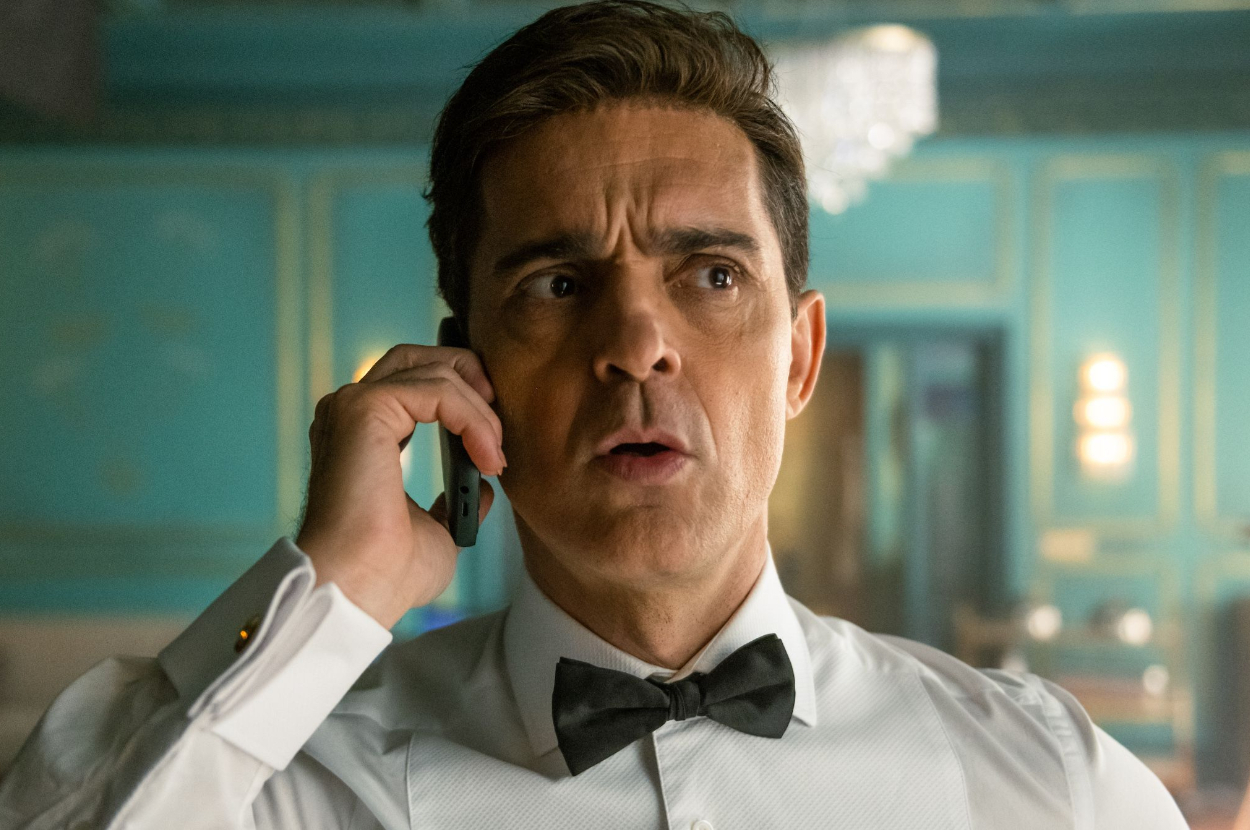 A man in a tuxedo with a bow tie speaks on a phone inside a luxurious room with elaborate decor