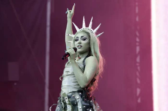 Lady Gaga performing on stage in a Statue of Liberty-inspired outfit with a spiked crown, raising one arm and holding a microphone