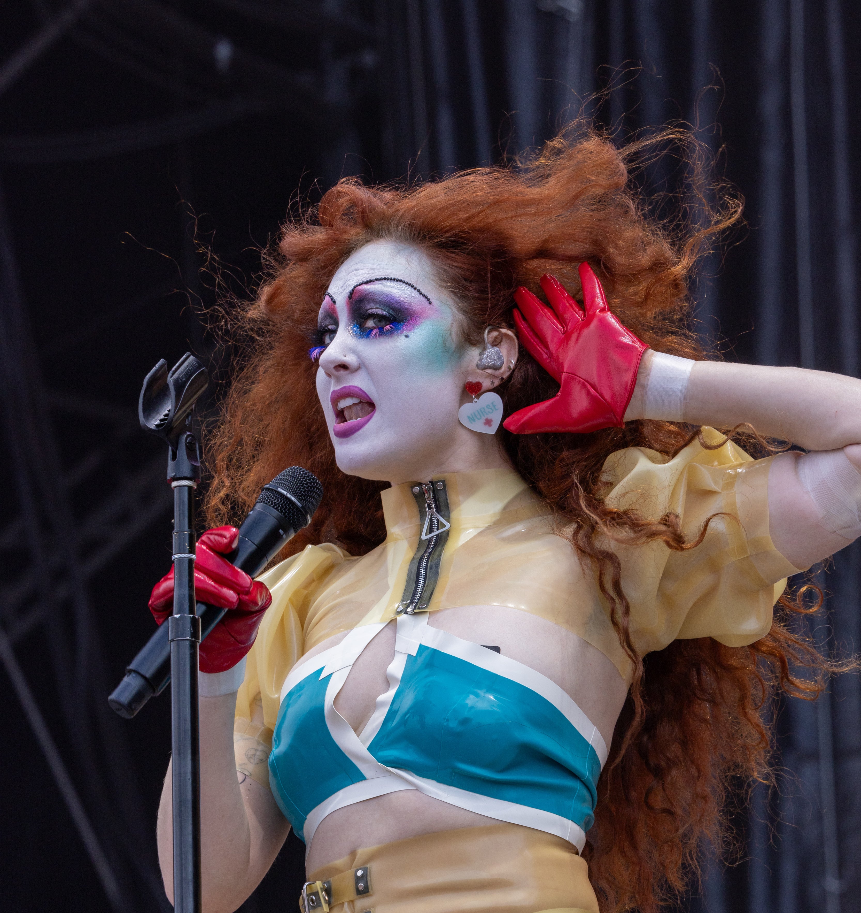 Performer with voluminous red hair wearing a vibrant, futuristic outfit sings on stage