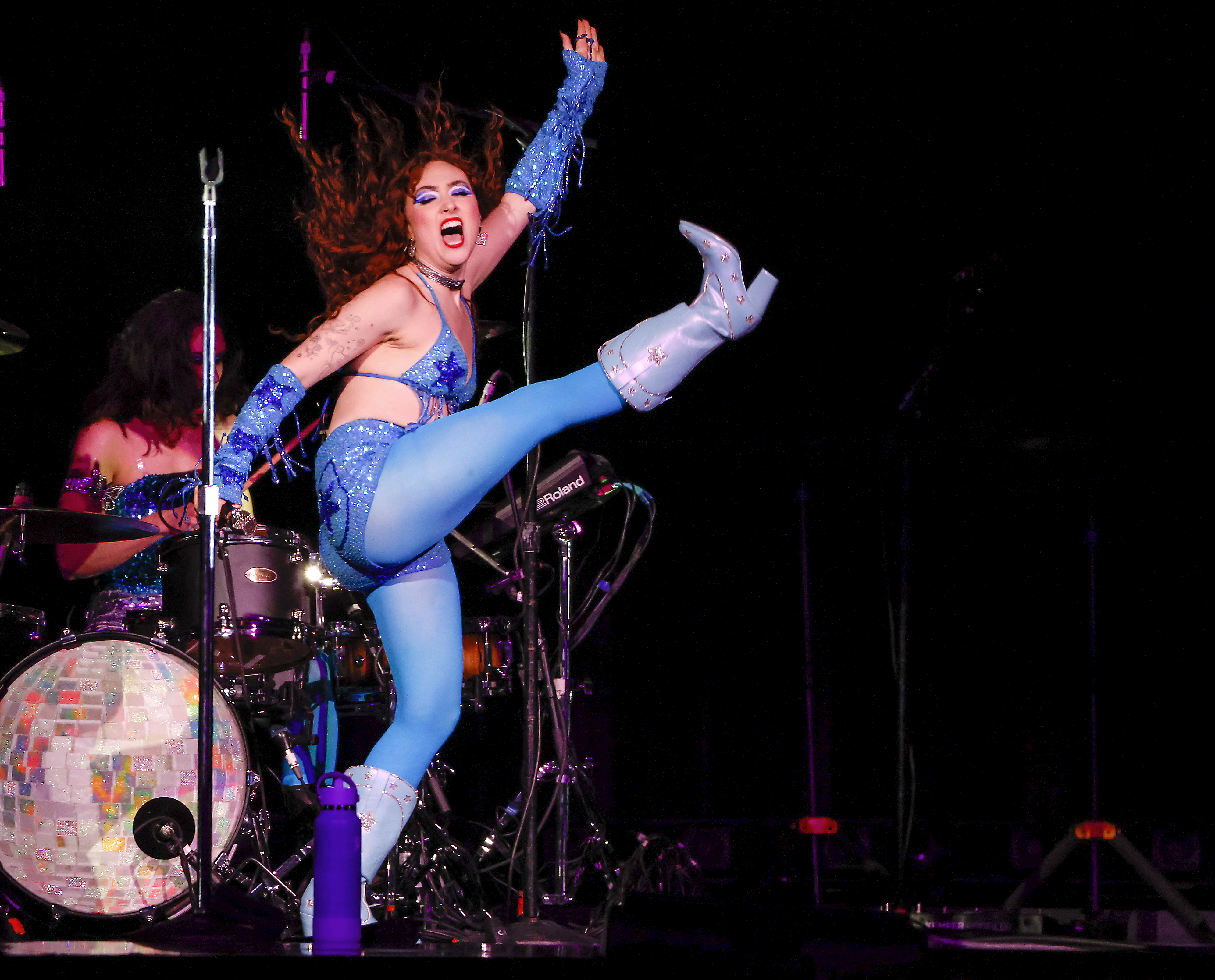 Lizzo performs energetically on stage, kicking her leg high, wearing a sparkly blue outfit and boots