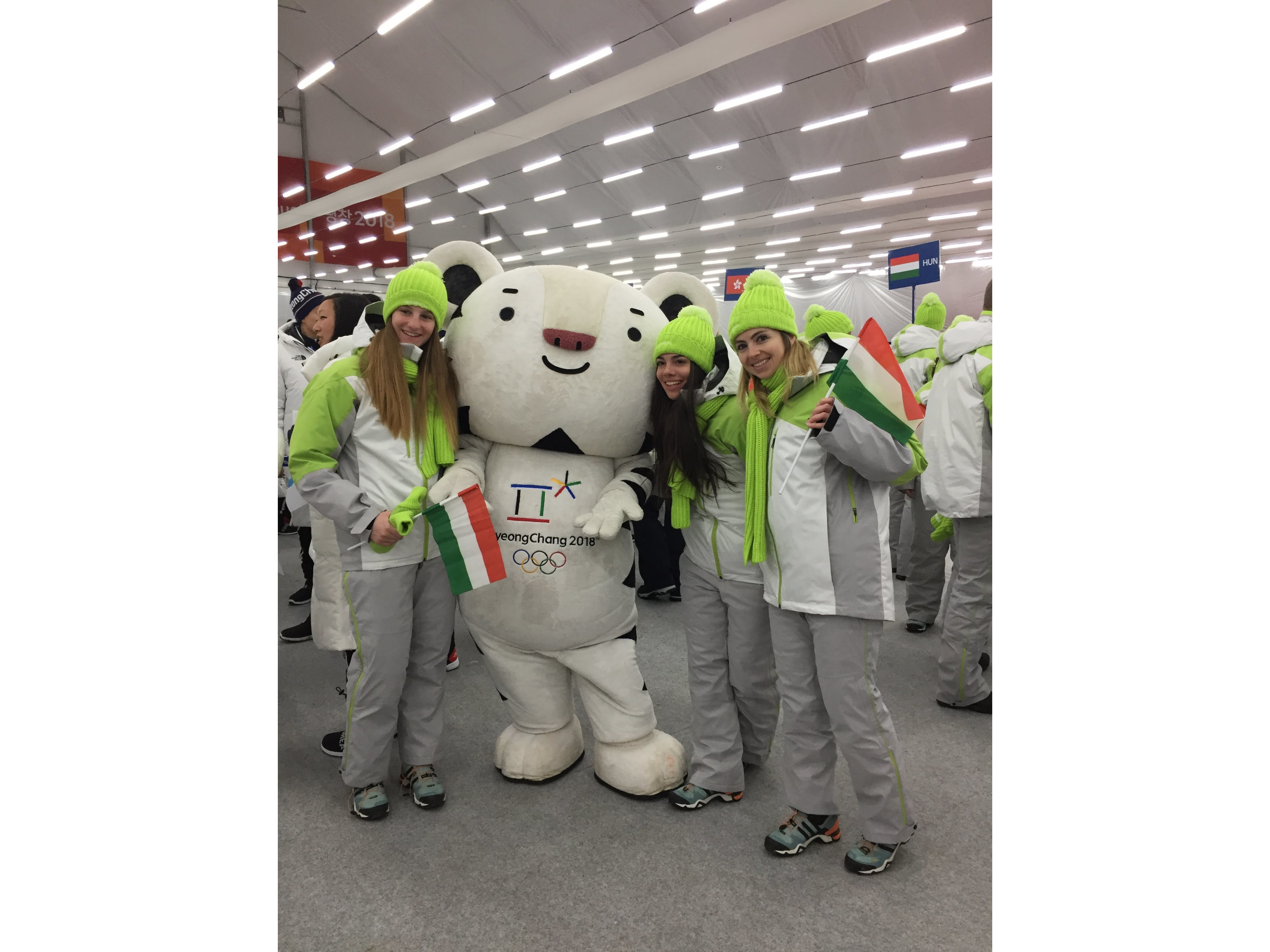 Mascot Soohorang at PyeongChang 2018 Winter Olympics poses with Elizabeth Swaney and two Hungarian Olympic teammates in winter jackets and hats holding flags. Other athletes and countries are in the background.