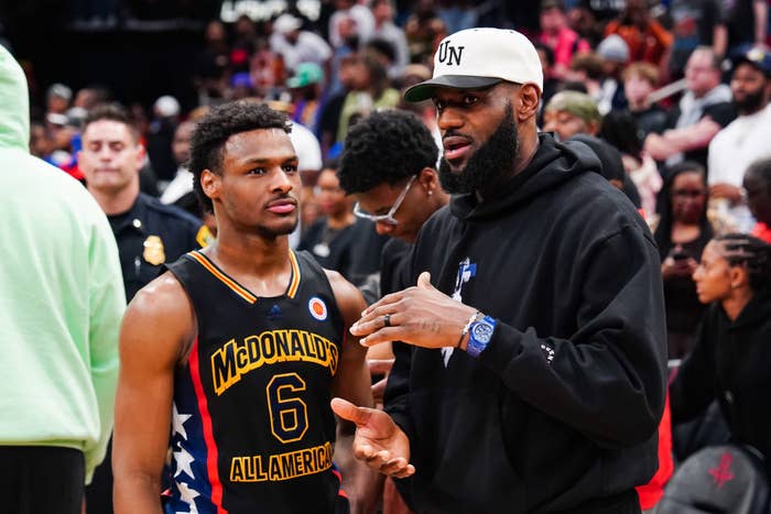 LeBron James in a black hoodie and hat talks to Bronny James in a black and yellow basketball jersey at a McDonald's All American event