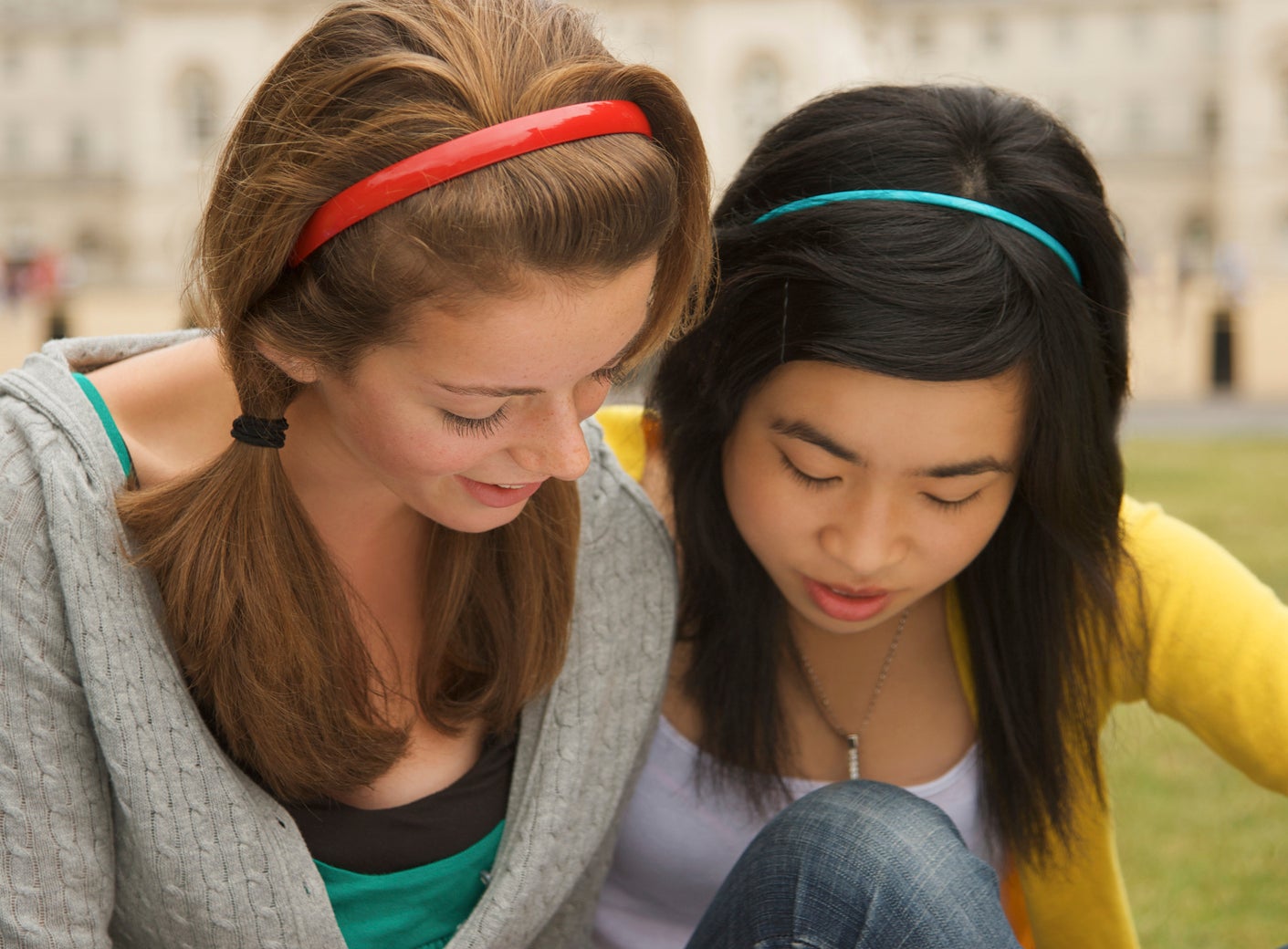 Two young women, one with a red headband and the other with a turquoise headband, sitting on a lawn, looking at a book together
