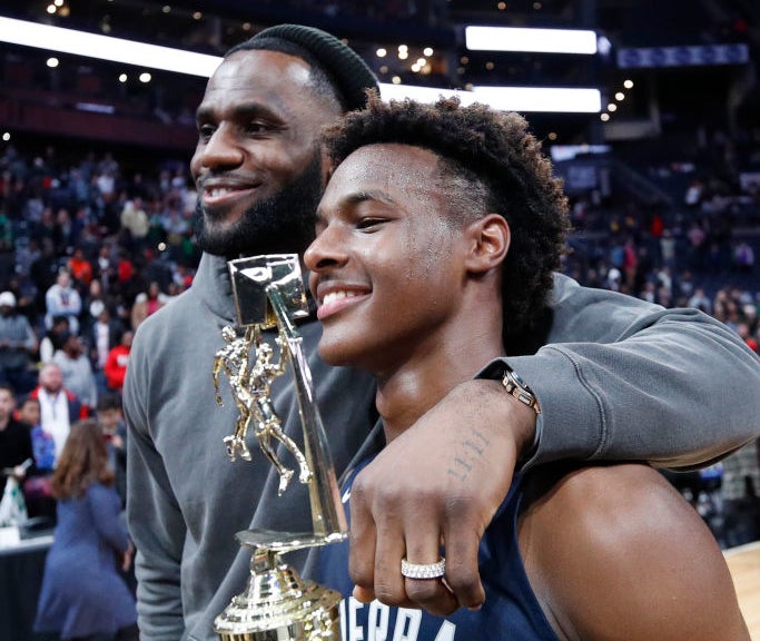 LeBron James poses with his arm around son Bronny James, who is holding a basketball trophy, on a basketball court
