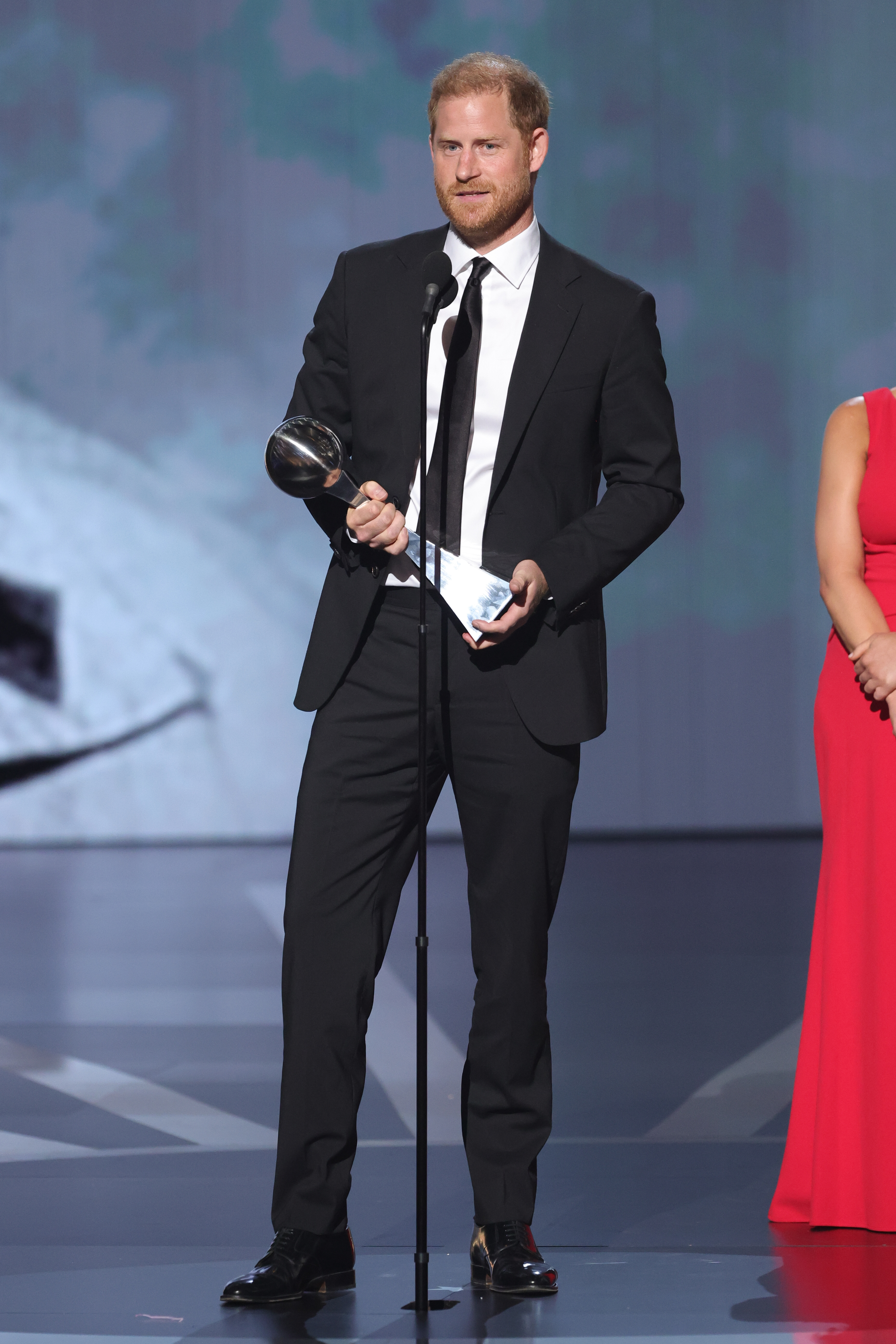 Prince Harry, dressed in a suit and tie, holds an award and speaks into a microphone on stage. An unidentified person in a gown stands nearby