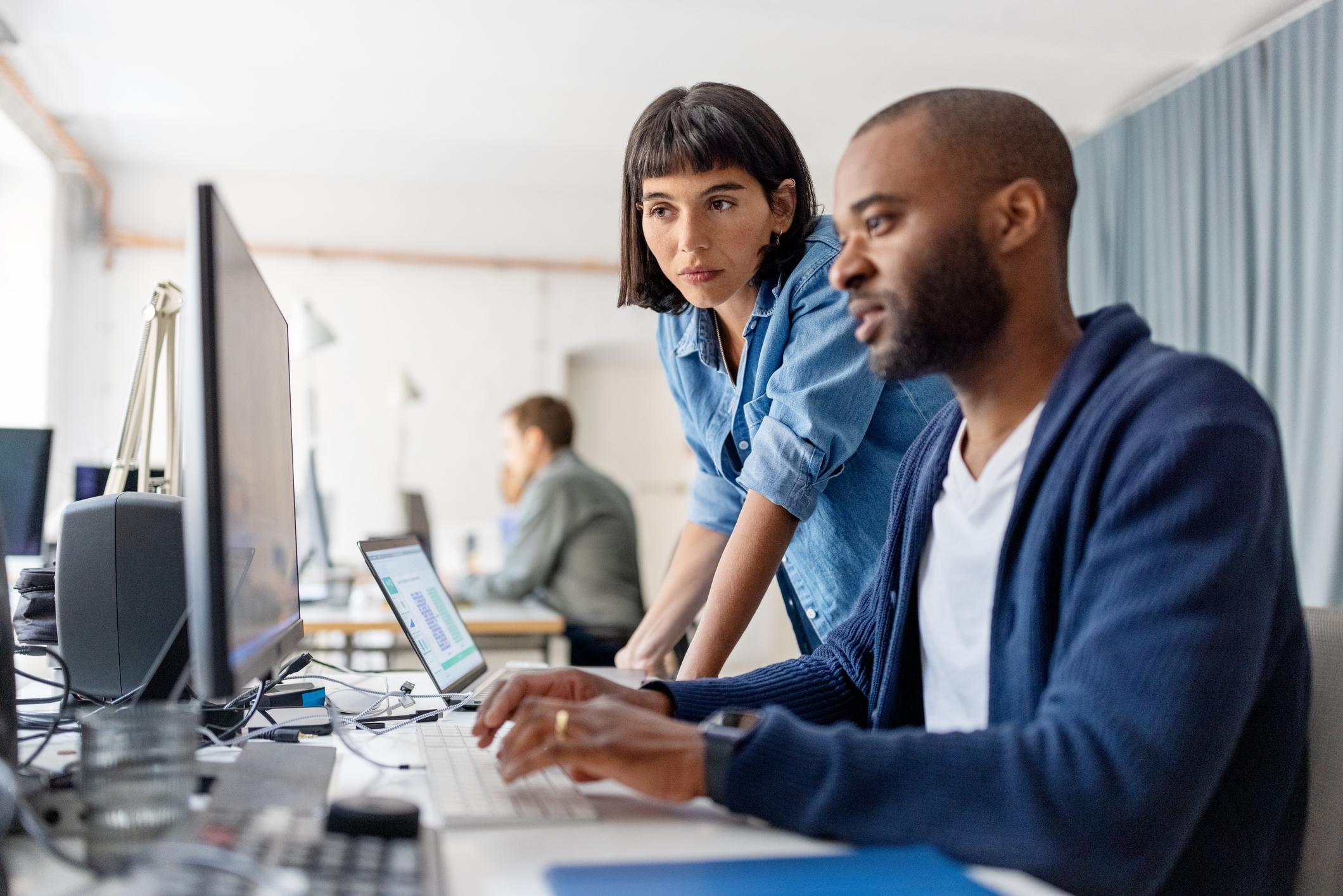 A woman and a man are working at a desk with computers. The woman is standing and looking at the man's screen. Another person is in the background