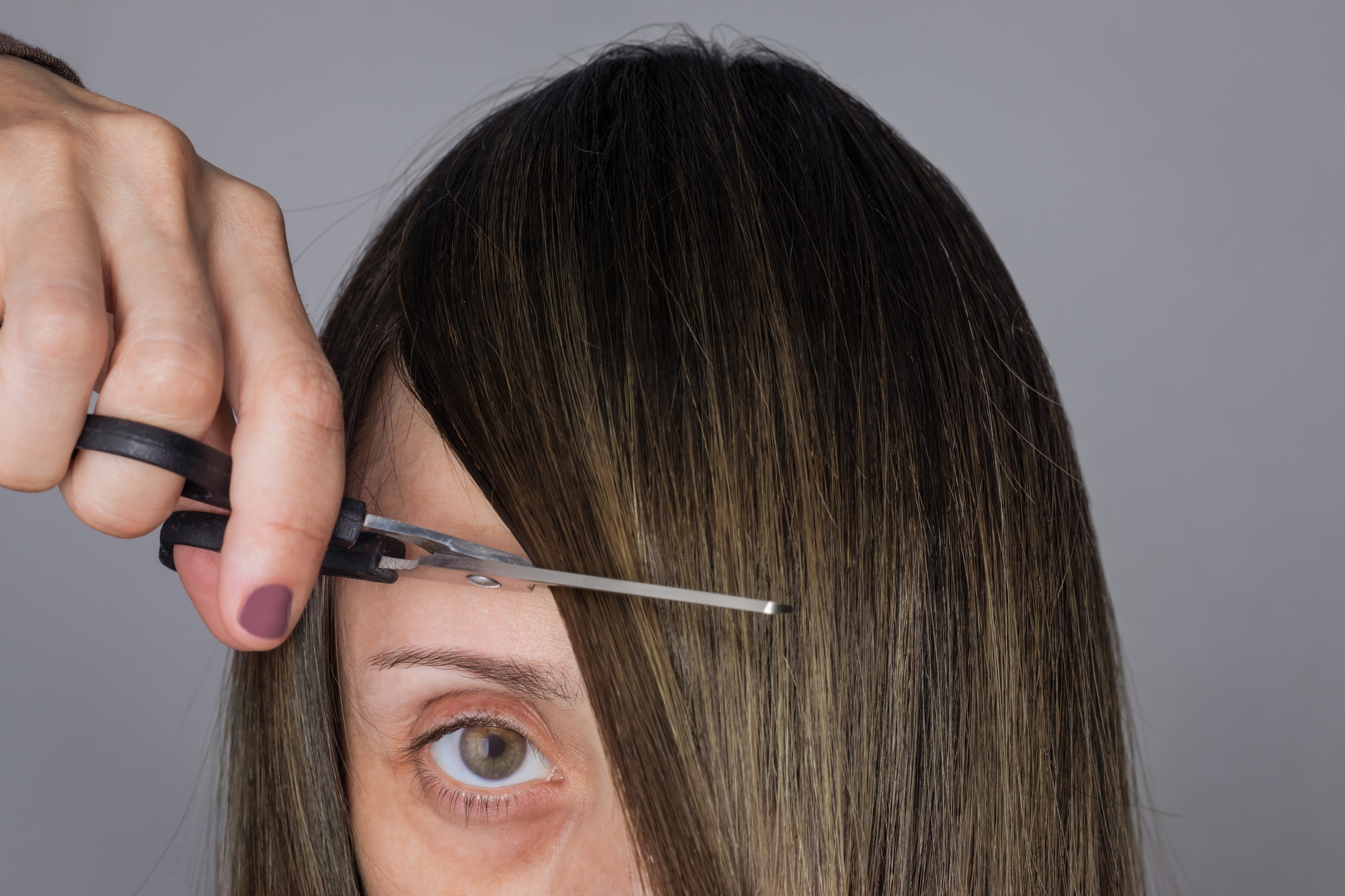 A close-up of a person cutting their own bangs with scissors. The person's eye and hand holding the scissors are visible
