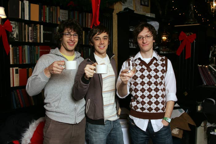 The Lonely Island in casual clothing hold up drinks and smile in a cozy, book-lined setting decorated for the holidays