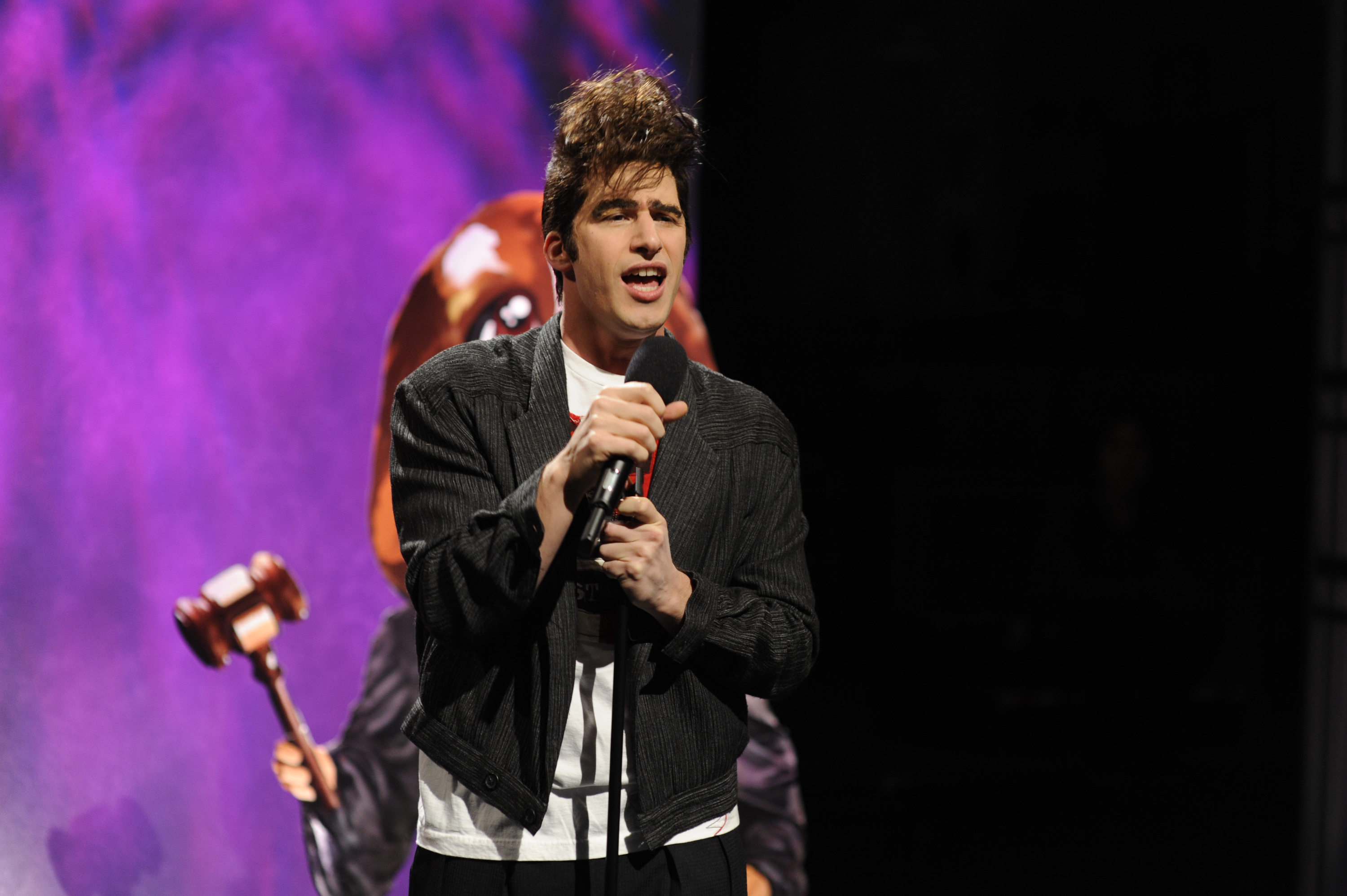 Andy Samberg performs on stage with a microphone, wearing a casual black jacket and a white t-shirt