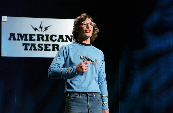 Andy Samberg in a comedic skit, holding a prop weapon in front of a sign that reads "American Taser." He is wearing a casual sweater and jeans