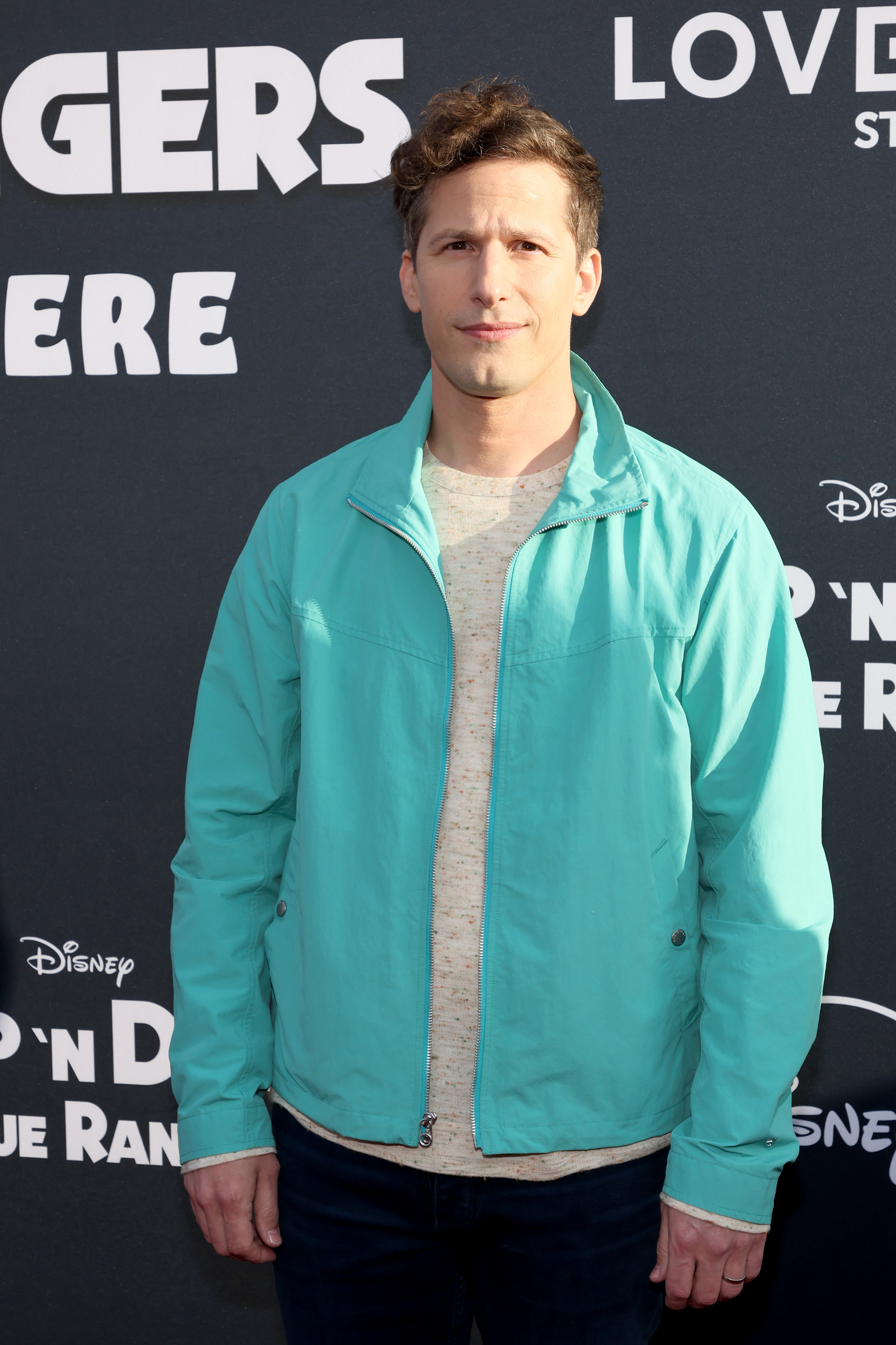 Andy Samberg on the red carpet, wearing a light jacket, long-sleeve shirt, and dark pants