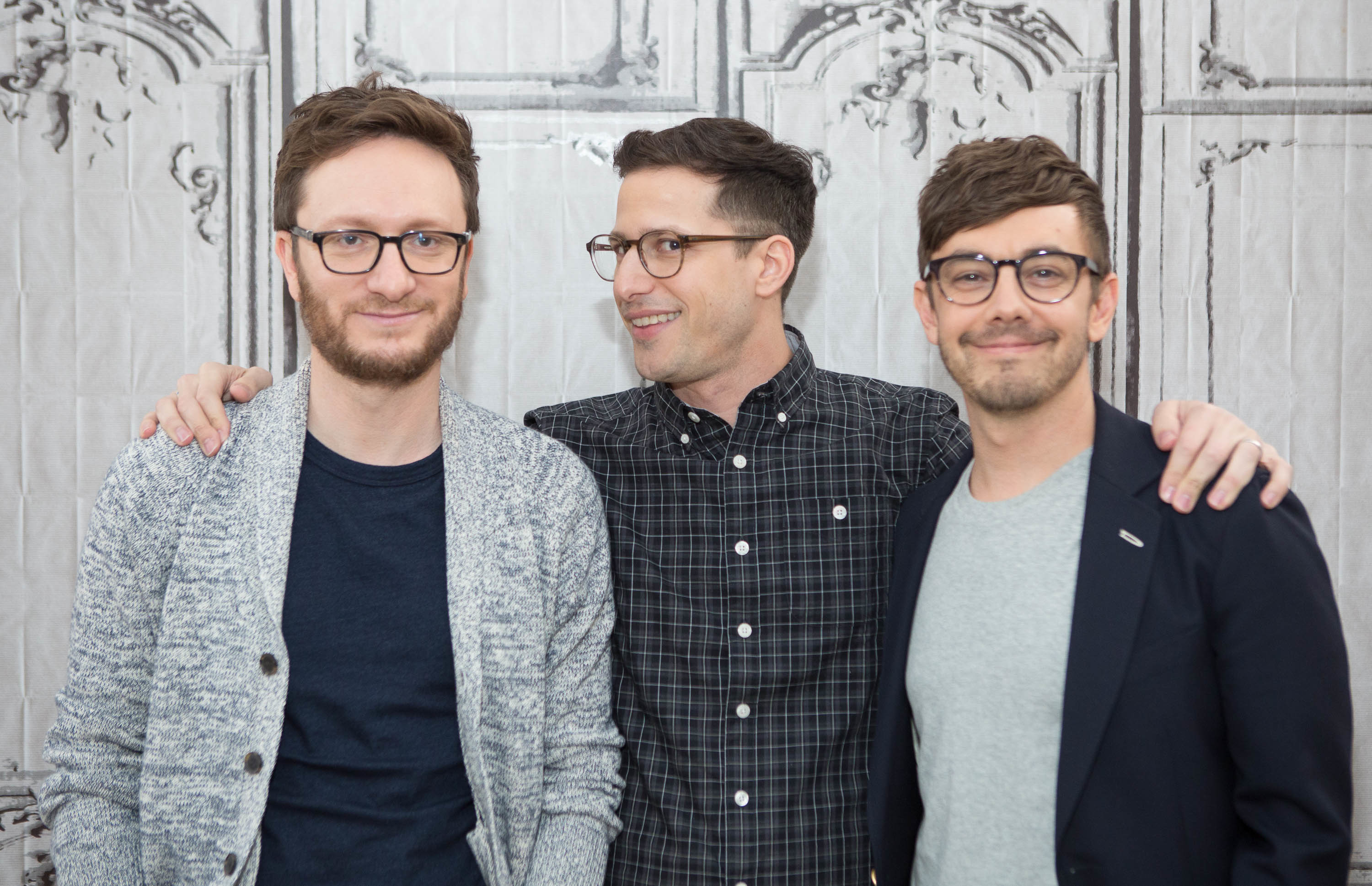 Jorma Taccone, Andy Samberg, and Akiva Schaffer are standing together, all wearing glasses. Jorma is in a cardigan, Andy in a checkered shirt, and Akiva in a blazer and t-shirt