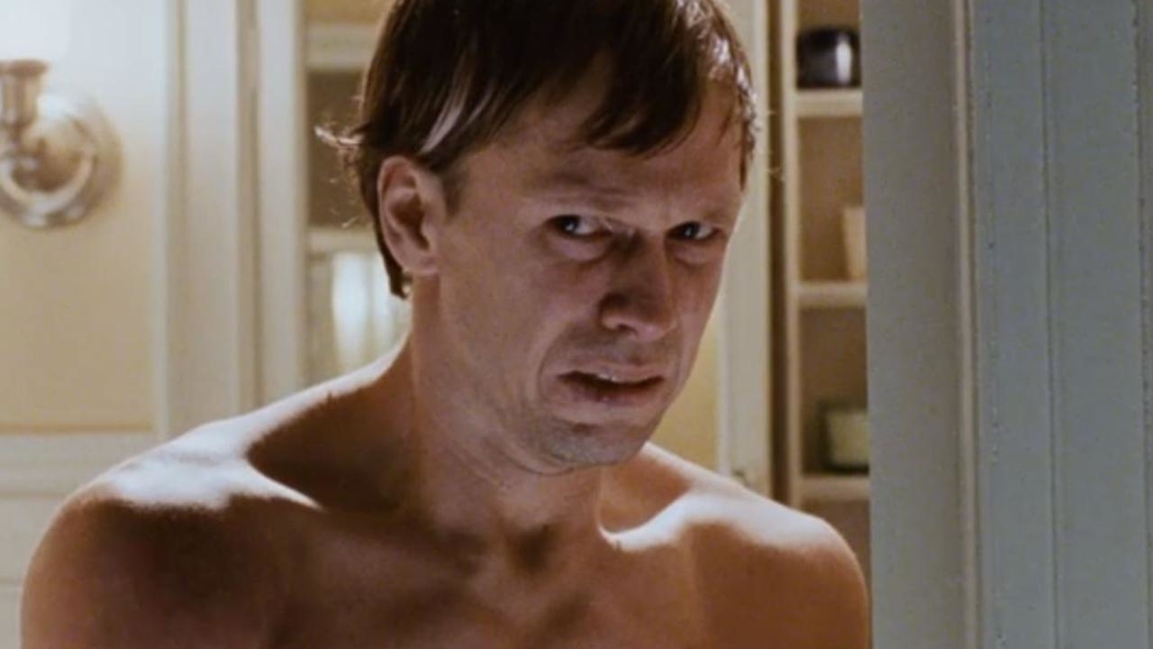 A man with a serious expression, shirtless, appears to be perplexed or anxious, standing indoors with a bathroom backdrop
