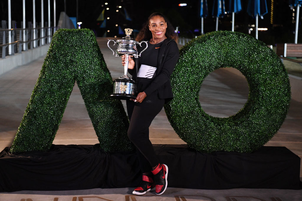 Serena Williams holds a championship trophy, standing in front of greenery arranged to spell "AO" on a dark backdrop. She wears a sporty outfit and smiles