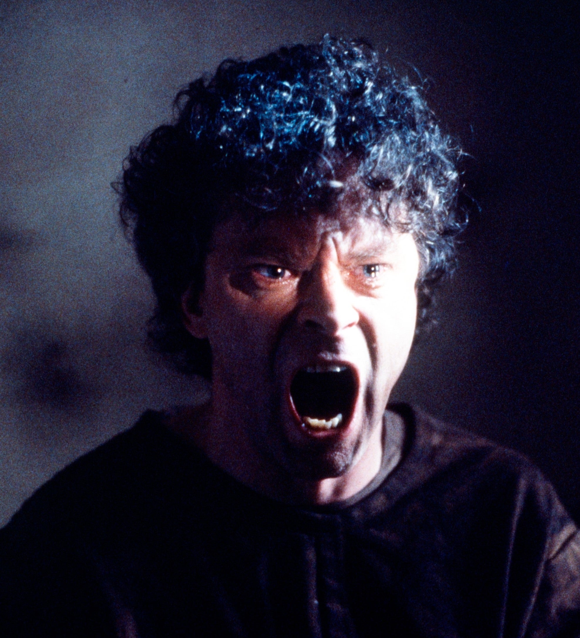 Man with curly hair screams with expression of intense anger or fear in a dramatic scene