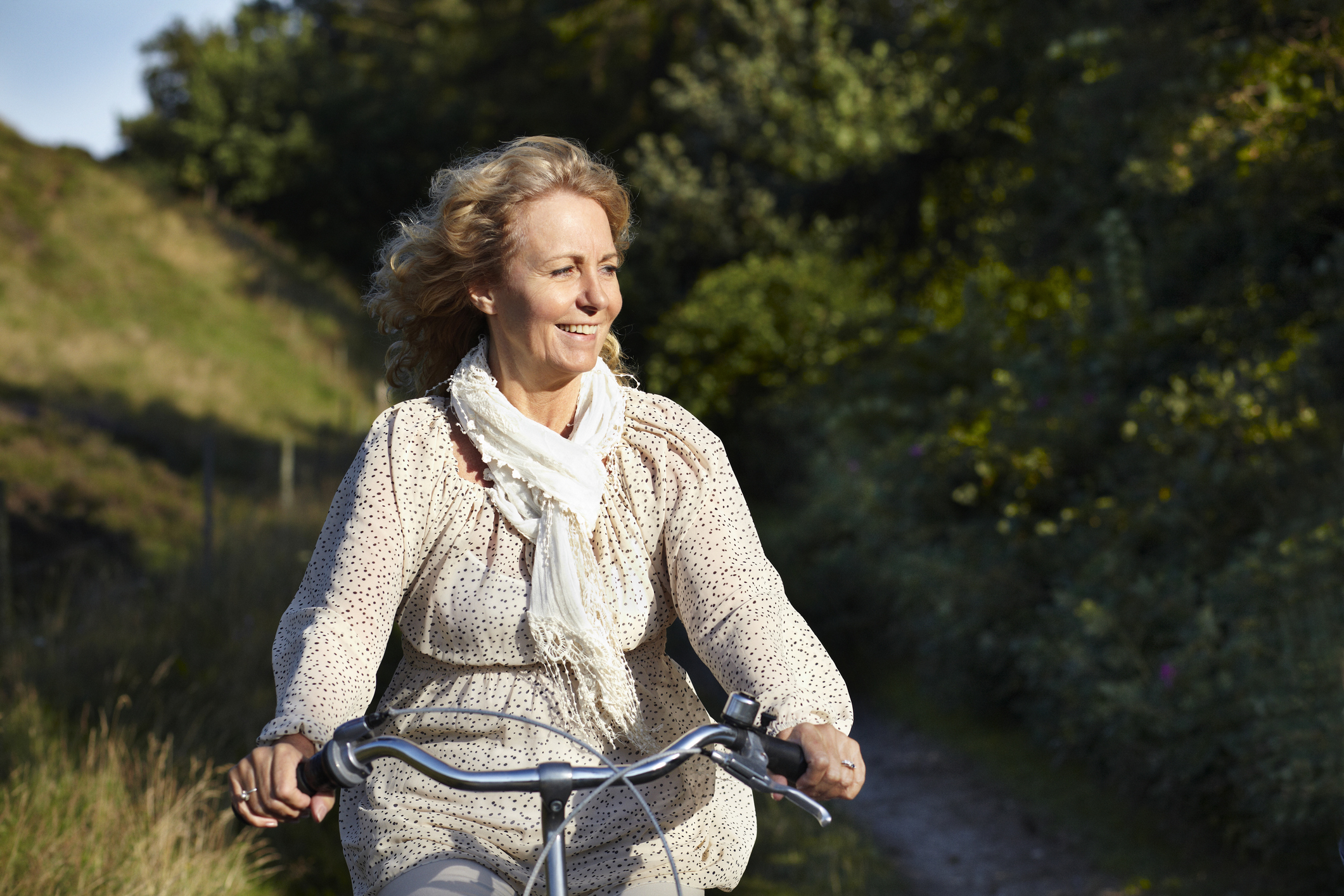A woman smiles while riding a bike on a forested trail, dressed in a flowy, patterned blouse and a light scarf